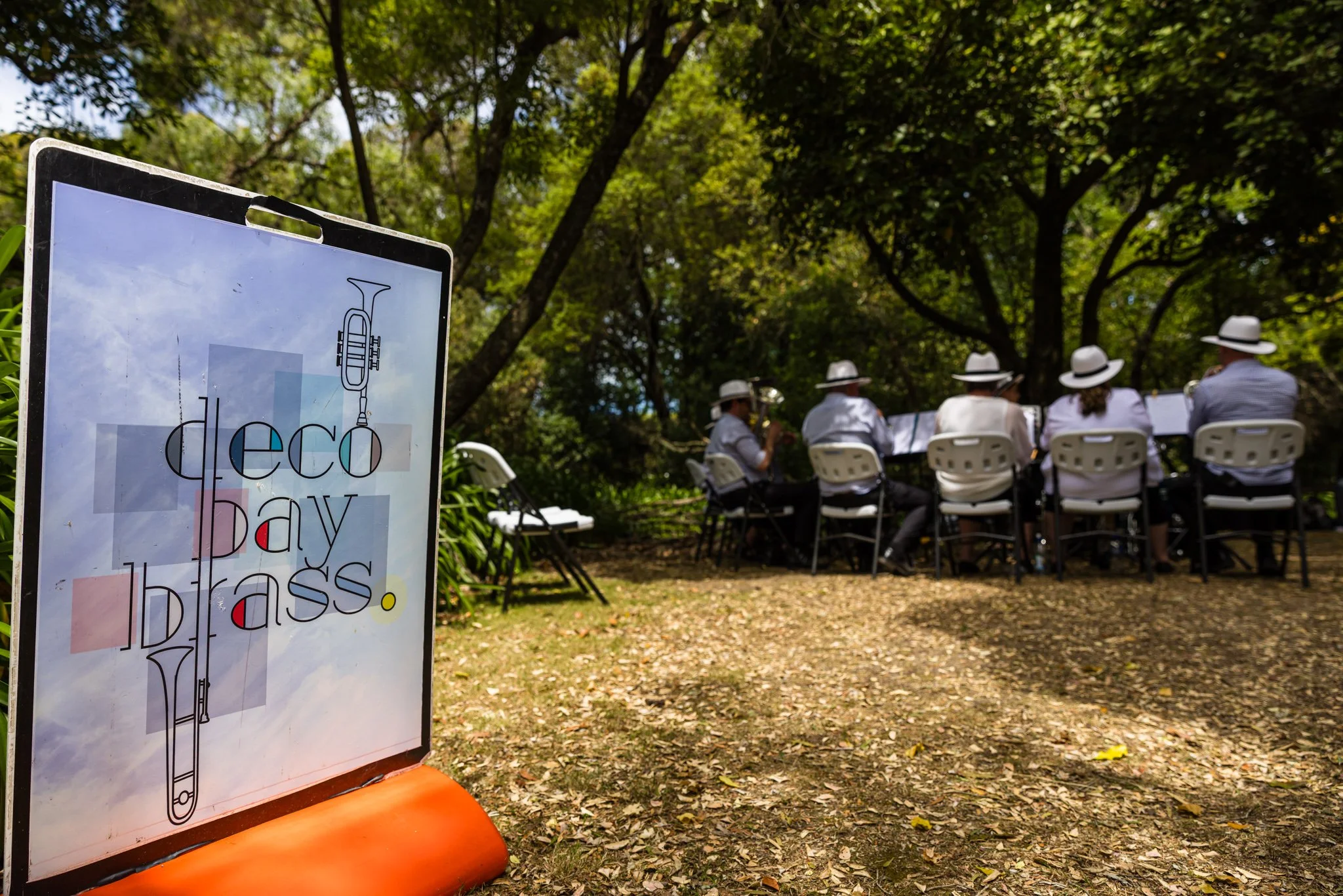 A sign with colorful abstract shapes and the words 'déco bay brass' next to a group of musicians playing brass instruments outdoors under trees.