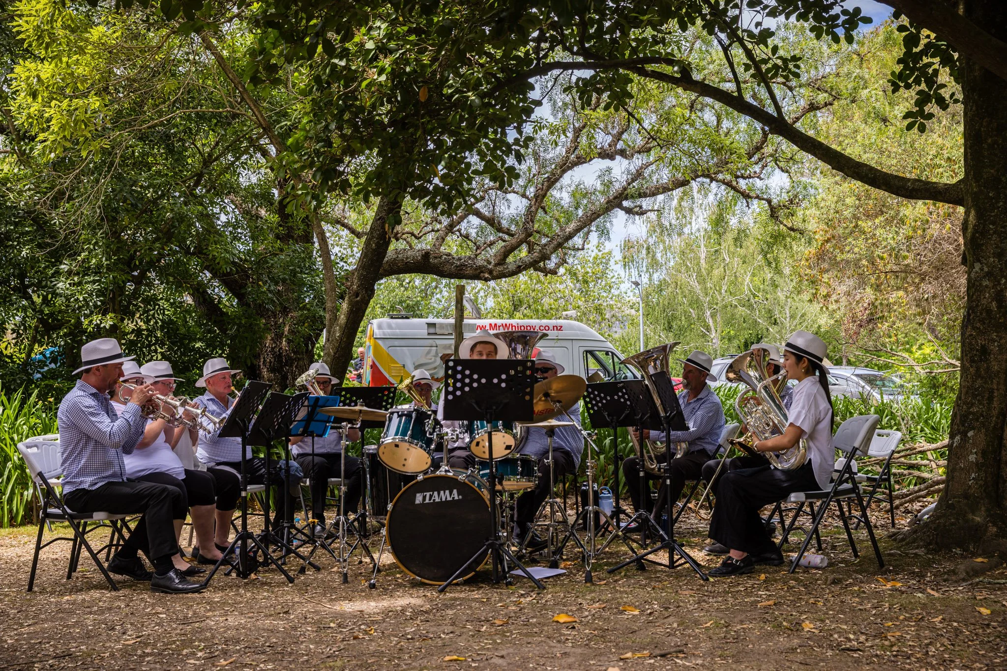 An outdoor band performing under a large tree, with musicians wearing white hats and playing various instruments such as trumpets, drums, and French horns, behind a music stand, with a vehicle and lush greenery in the background.