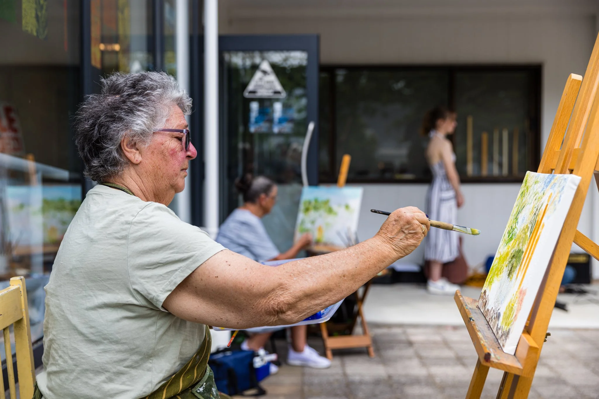 Older woman painting outdoors on an easel with two other women also painting in the background.