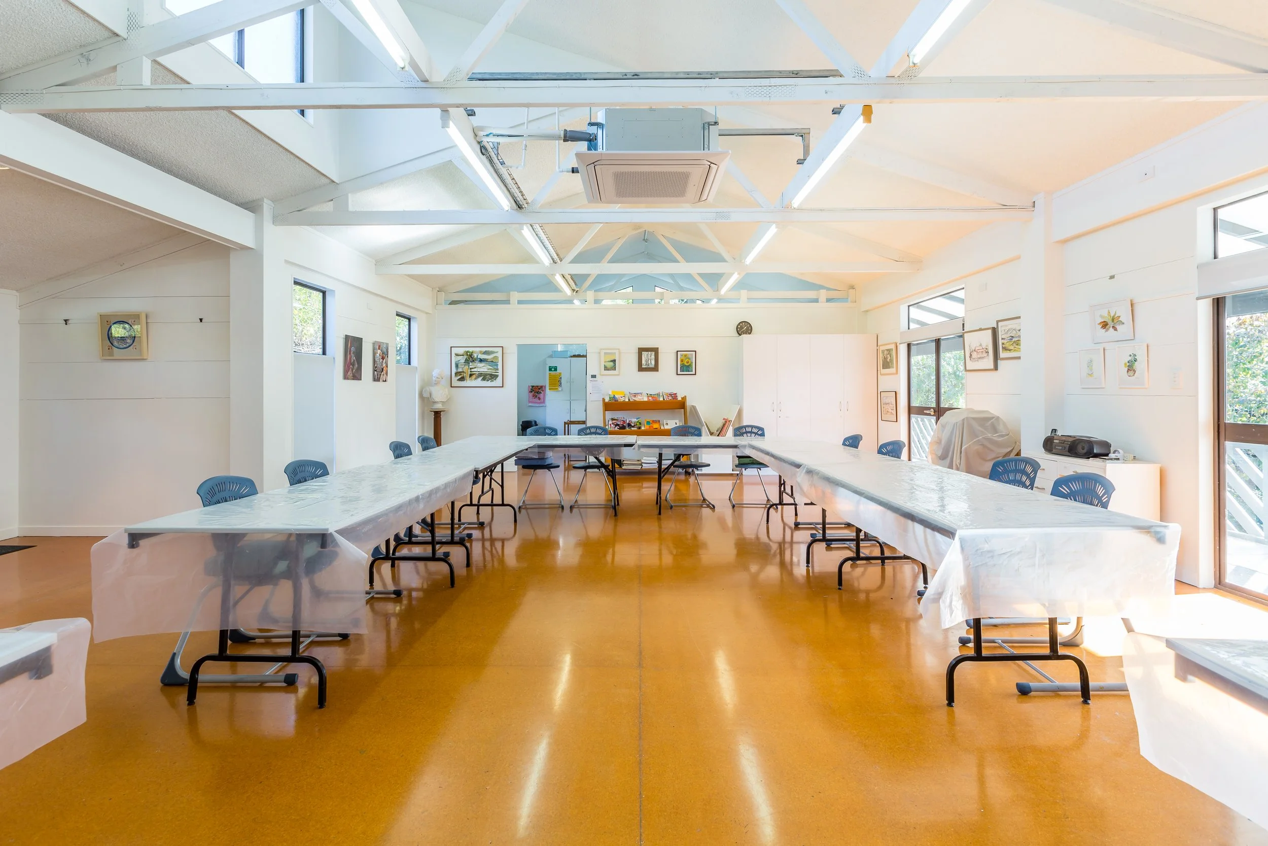A spacious conference room with long tables covered with white tablecloths, surrounded by blue chairs, and decorated with framed artwork on white walls. Large windows let in natural light.