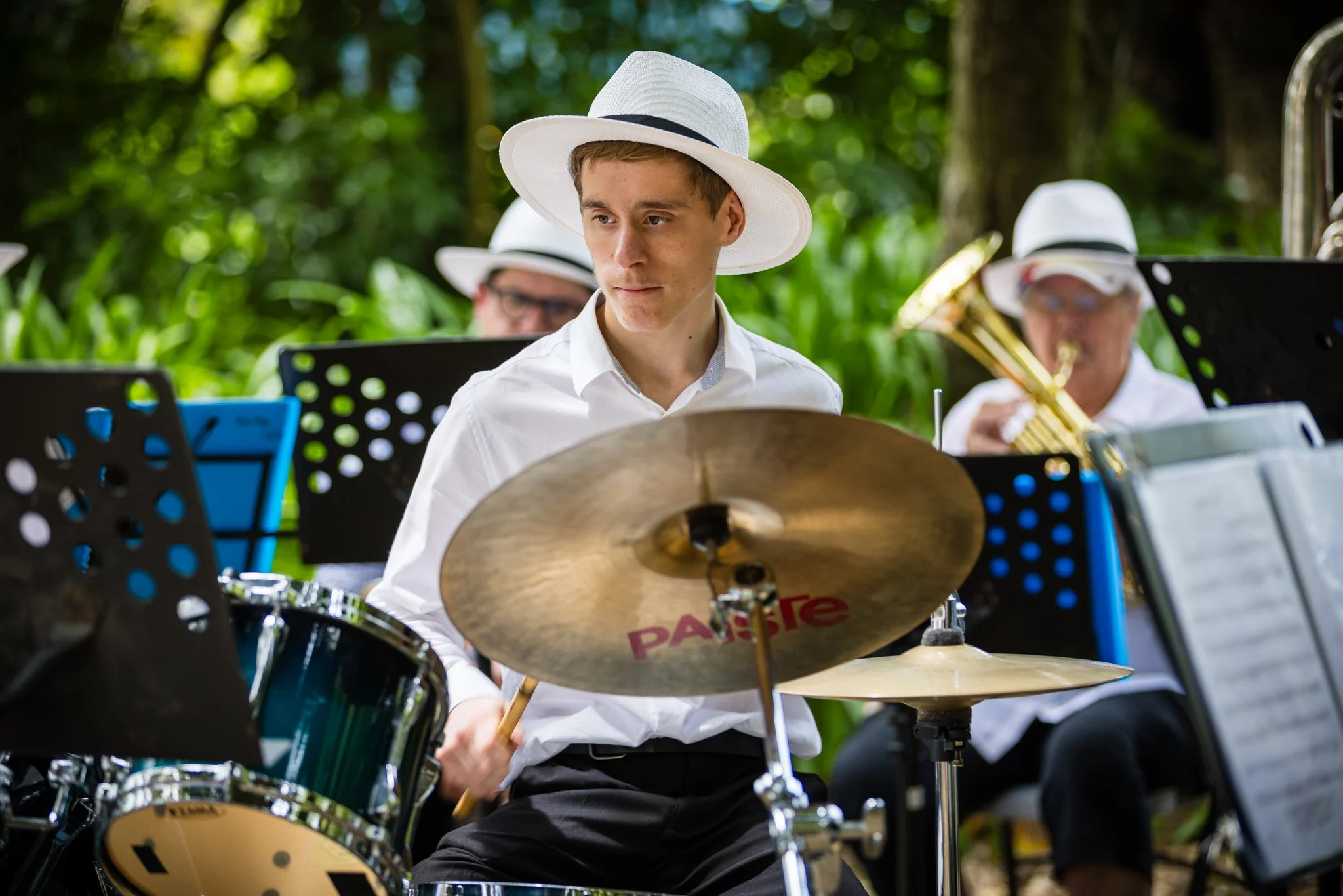Young man playing drums at an outdoor musical performance in a park, wearing a white hat and white shirt, with band members playing brass instruments in the background.