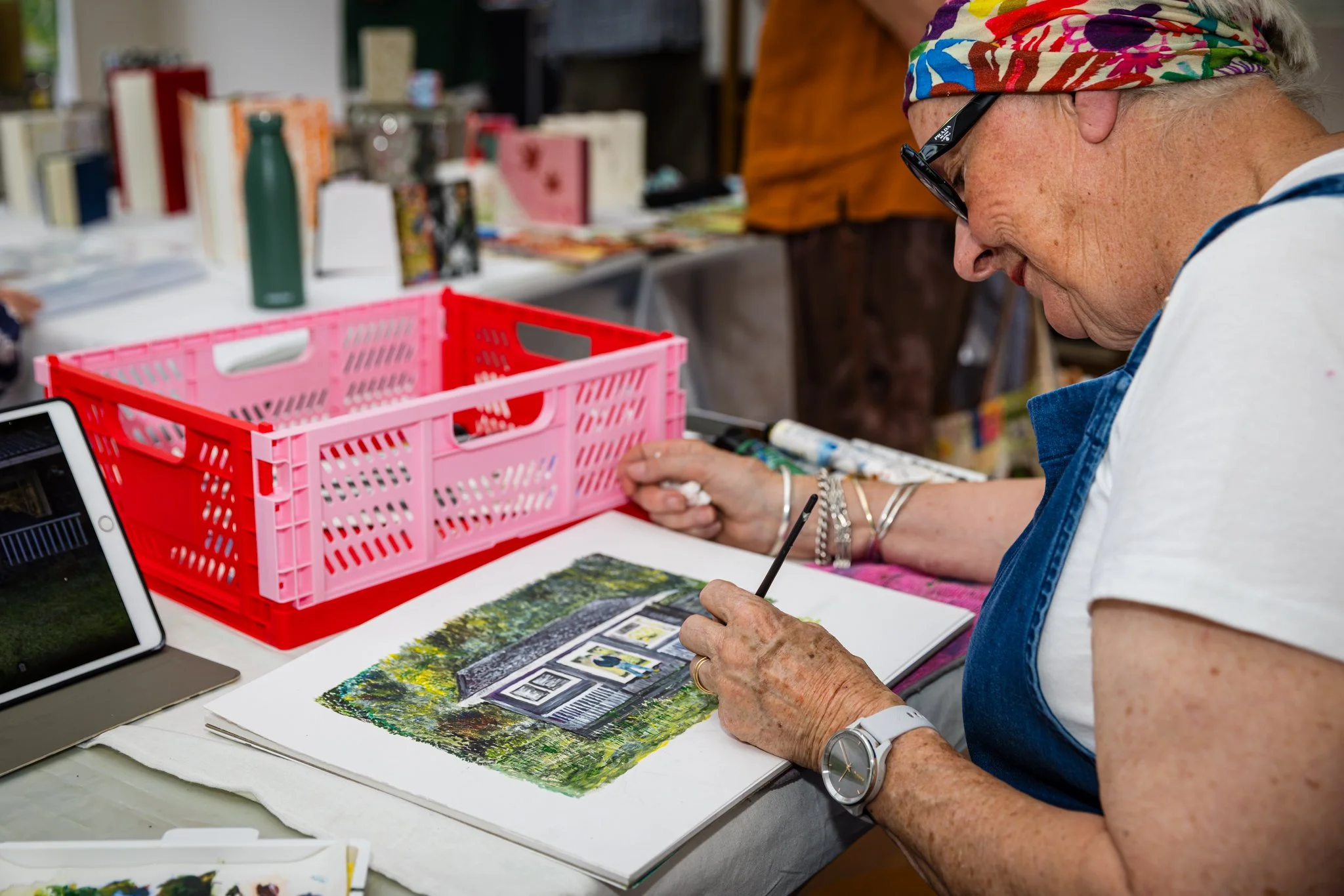 An elderly woman with a colorful headscarf, sunglasses, and multiple bracelets is painting a detailed scene of a house with trees on paper at a table. There is a tablet and a pink plastic basket on the table, along with various art supplies.
