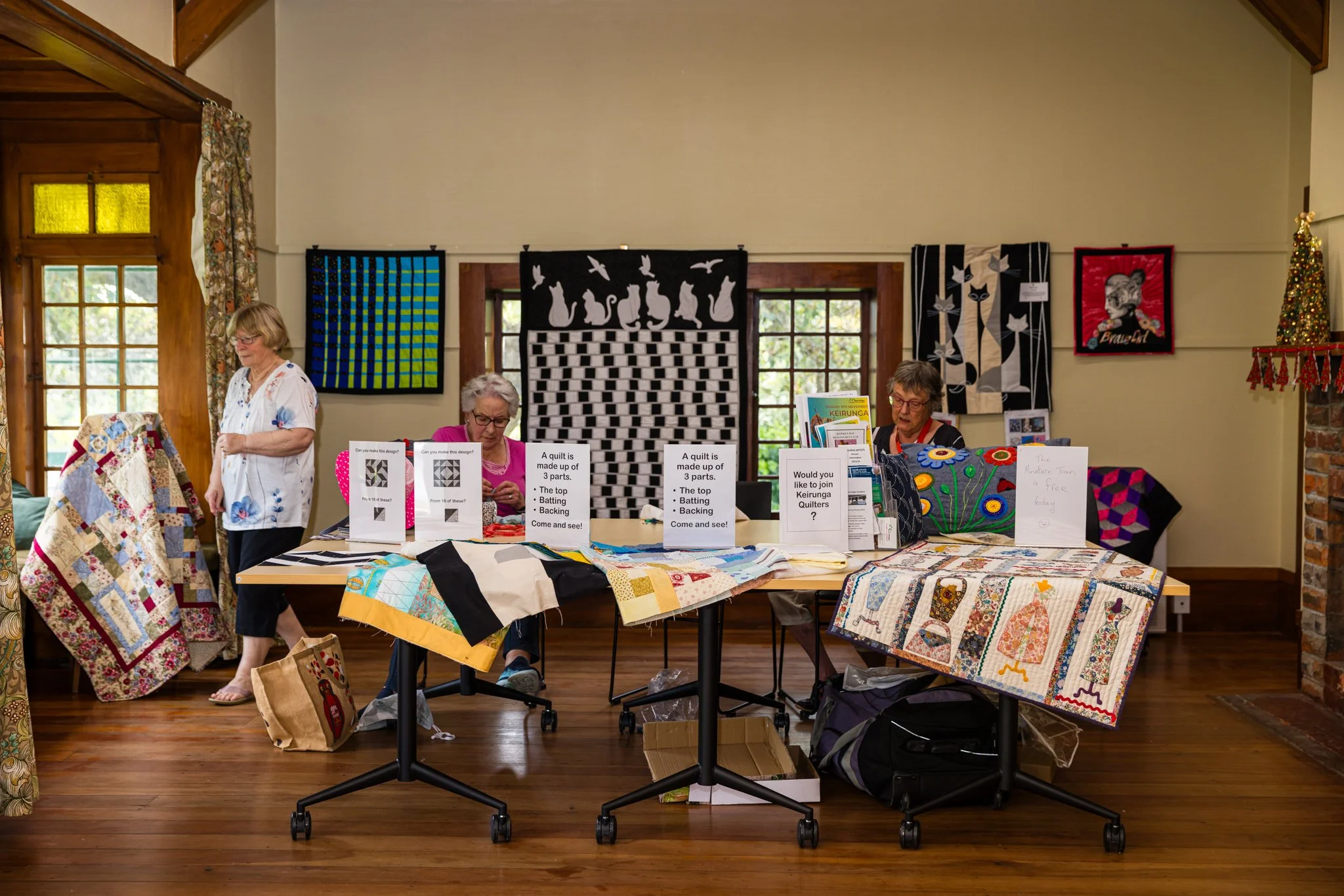Three women and one man at a booth displaying quilted blankets, with colorful quilt wall hangings behind them.