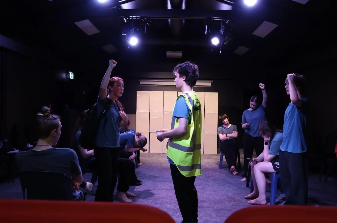 Group of people in a theater or rehearsal space, with some sitting and some standing, engaging in a discussion or activity, lit by overhead stage lights.