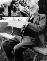 An elderly man with glasses sitting on a park bench, holding a cane and reading a newspaper.