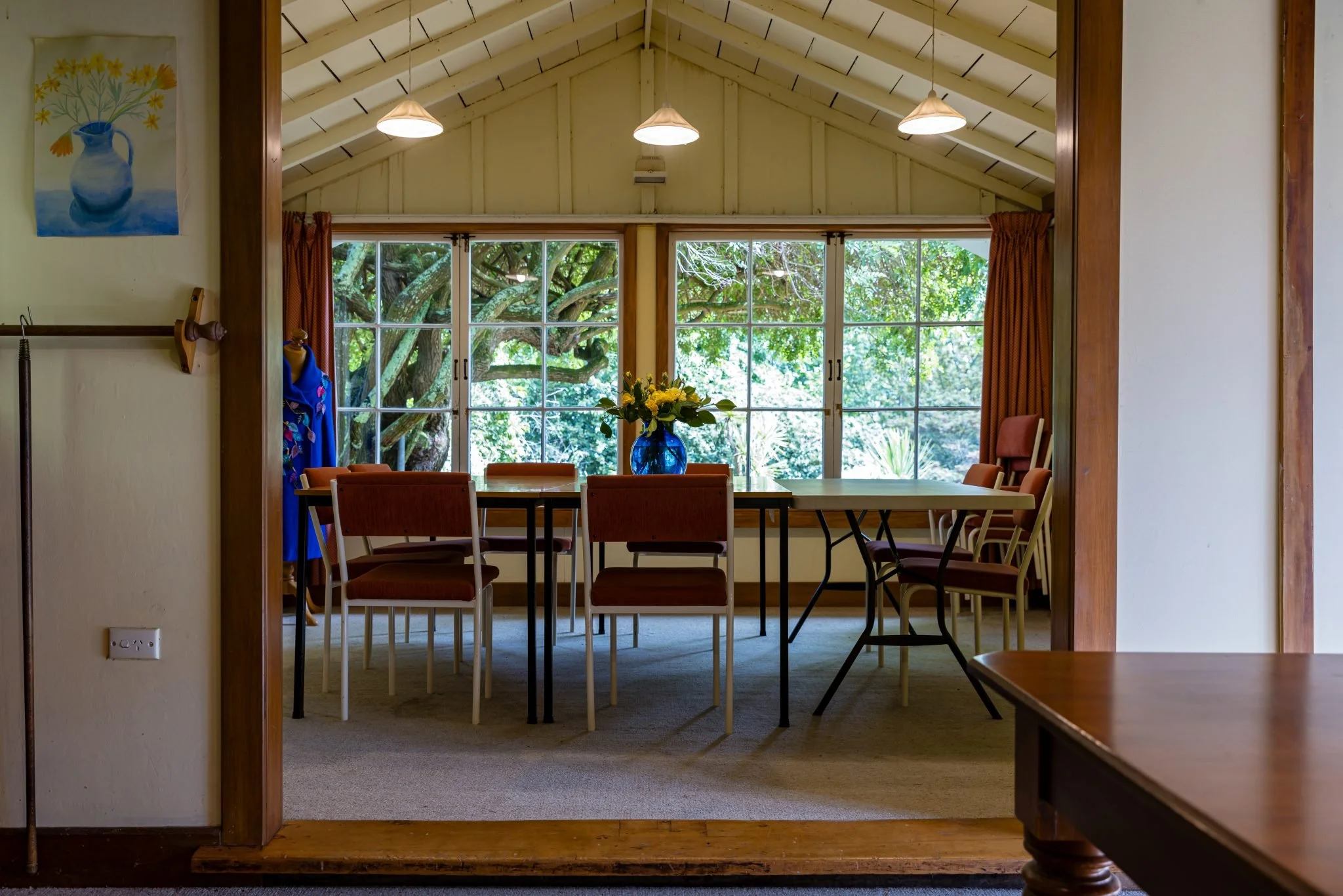 A dining room with a large window showing green trees outside. There are 8 chairs around a rectangular table with a blue vase of yellow flowers on top. The ceiling is vaulted with hanging lights.