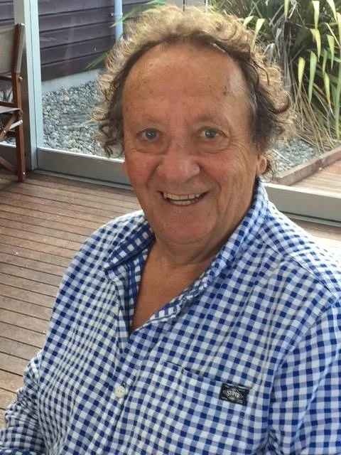 Smiling man with curly hair wearing a blue and white checkered shirt, inside a room with wooden floor and large window showing outdoor plants.