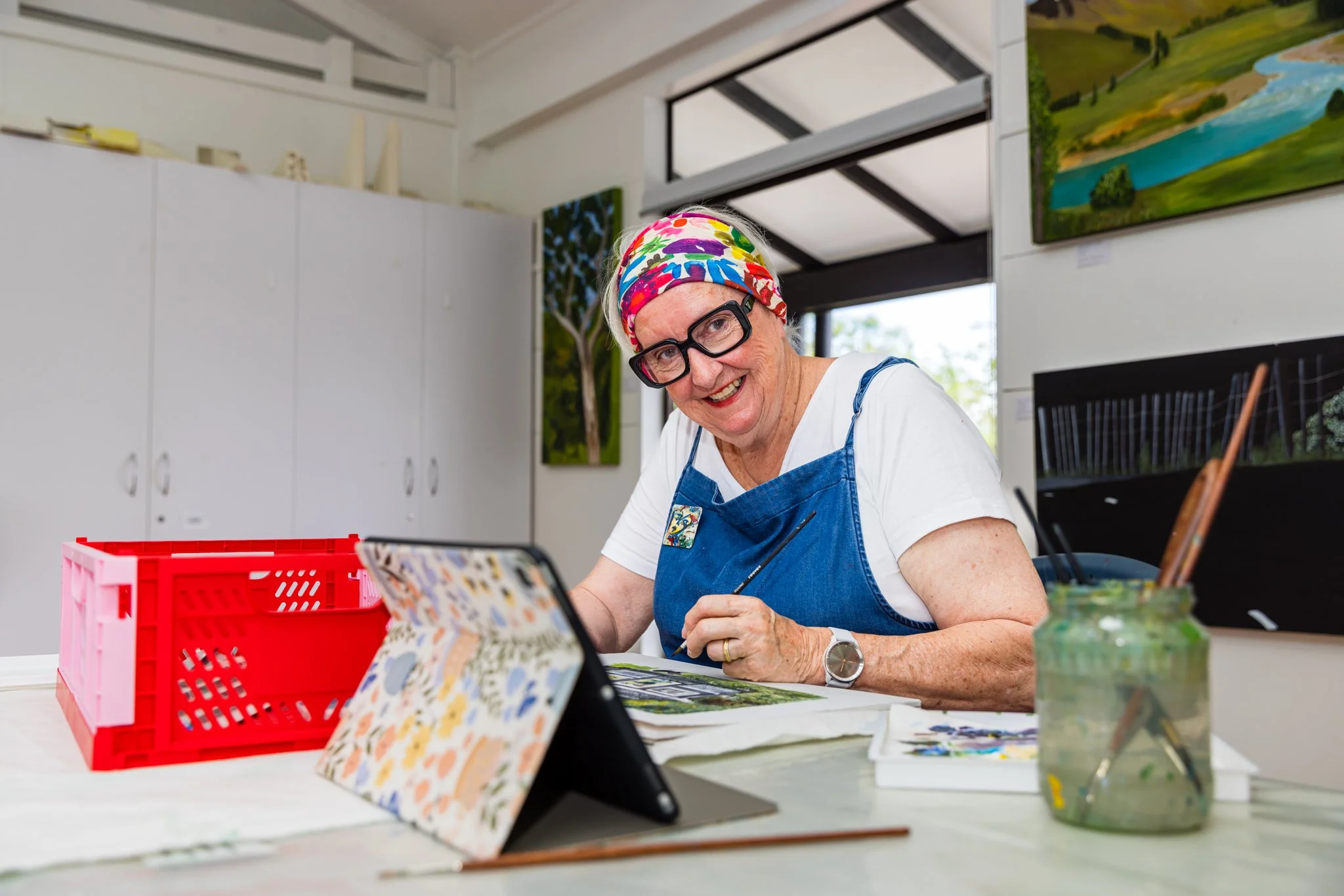 An elderly woman wearing glasses, a colorful headscarf, and a denim apron, smiling and working on an art project at a table with various art supplies, in an art studio with paintings hanging on the walls.