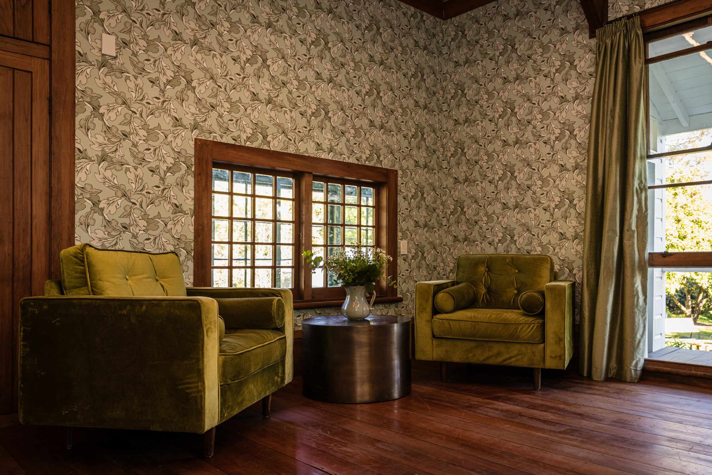 Living room with two green velvet armchairs, a dark round coffee table with a white vase and flowers, wood paneling, floral wallpaper, and large windows letting in natural light.