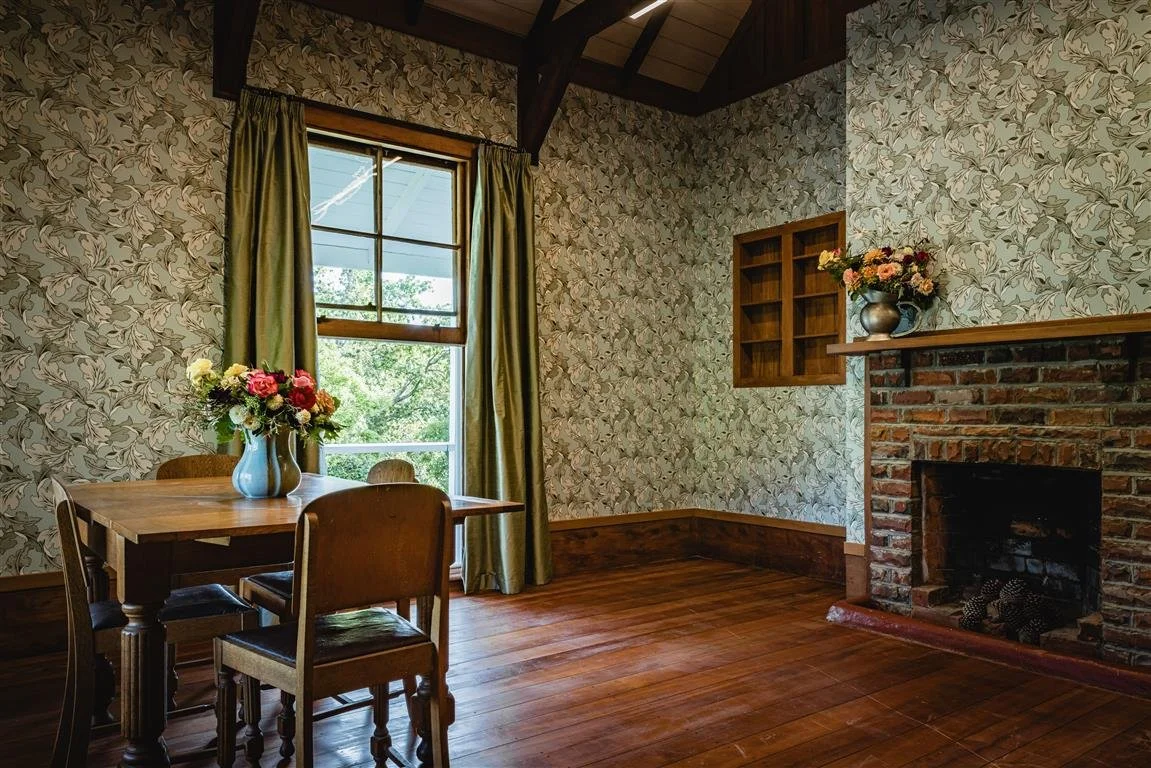 A cozy dining area with a wooden table and chairs, a window with curtains, floral wallpaper, a brick fireplace with a vase of flowers on the mantel, and wooden flooring.