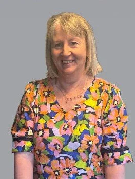 Smiling woman with blonde hair wearing a colorful floral dress, standing against a plain gray background.