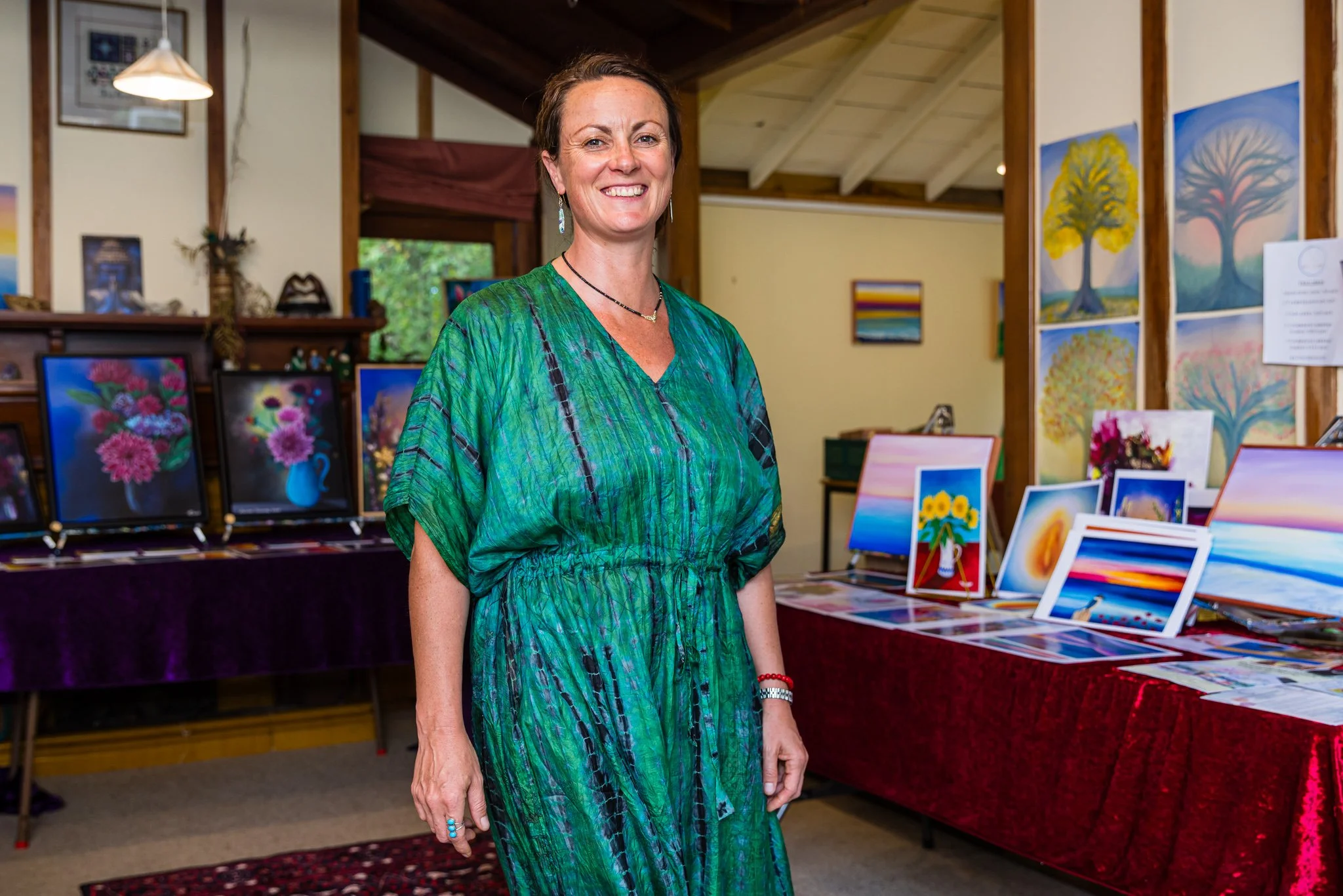 A woman standing in an art gallery with colorful landscape and floral paintings on tables and walls behind her.