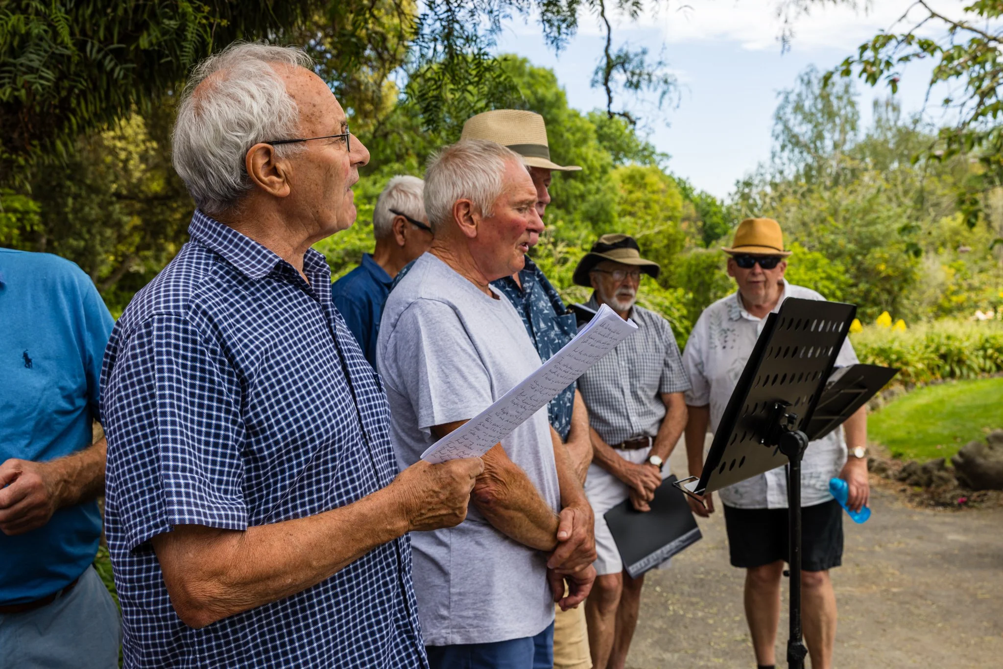 Group of elderly men singing outdoors in a garden, some holding sheet music, with lush green trees and plants in the background.