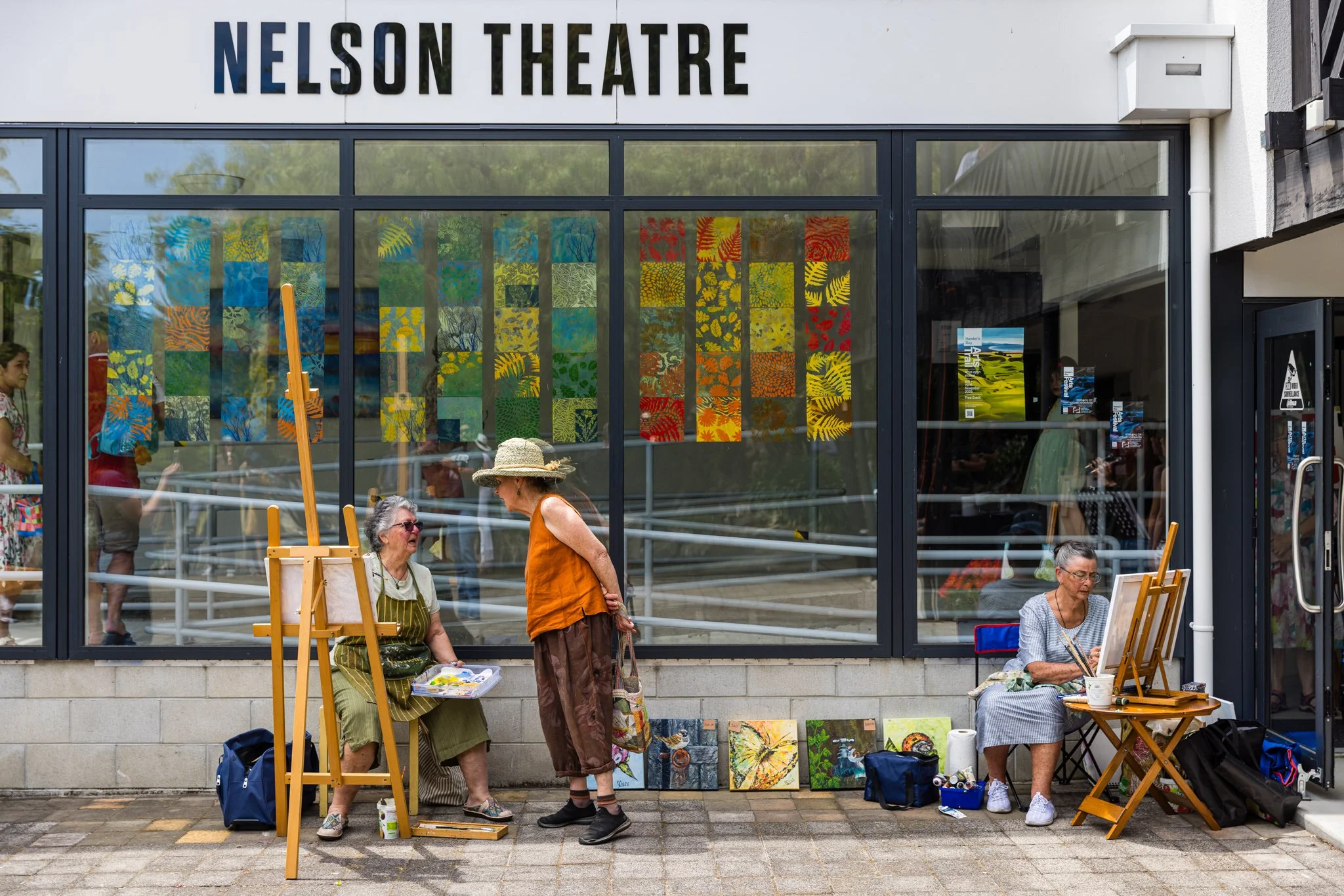 Art vendors set up outside Nelson Theatre with paintings on display and artists painting, in front of large glass window showing colorful artwork inside, on a sidewalk with people walking by.