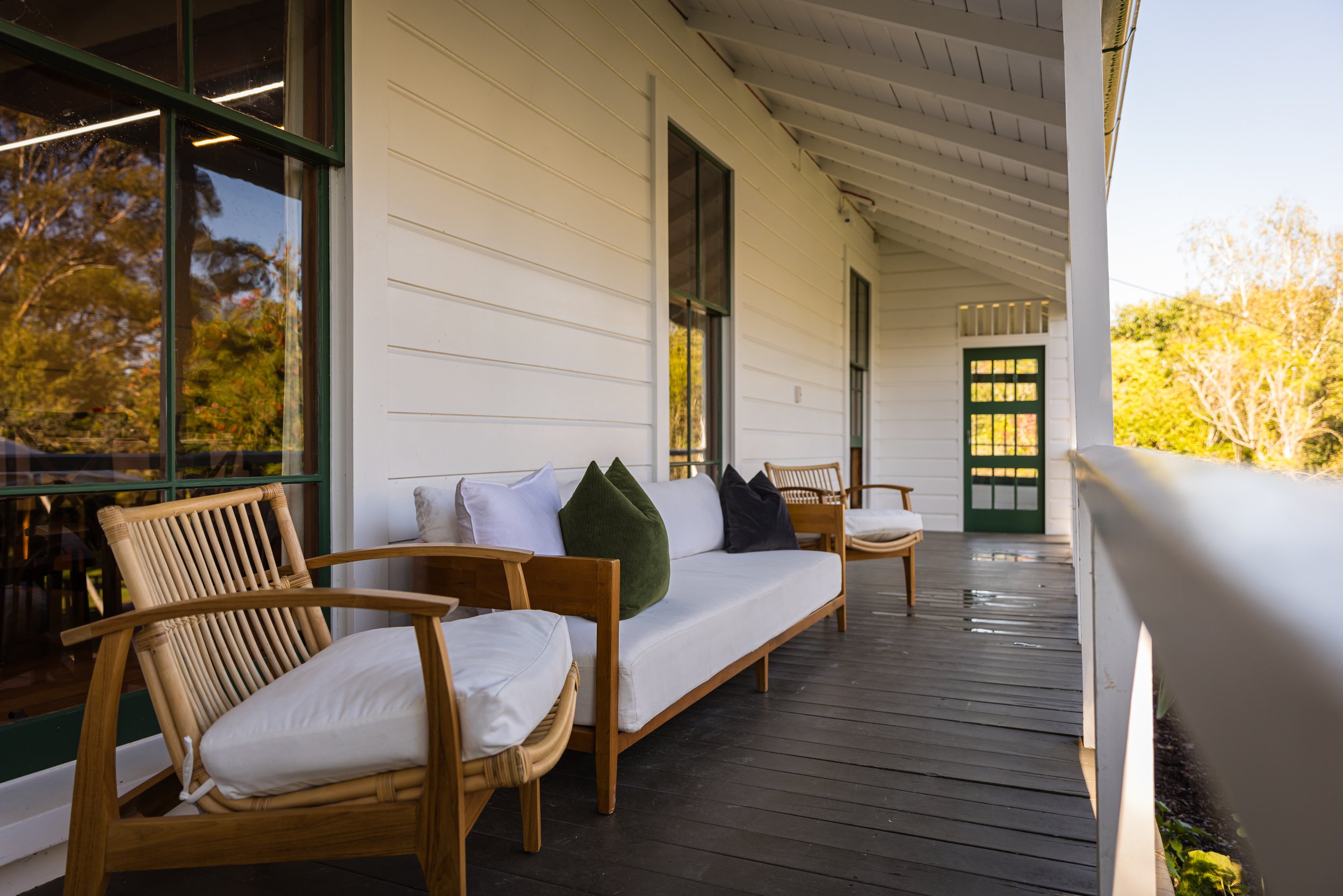 Front porch with white wooden siding, a green front door with glass panes, a black screen door, and wooden outdoor furniture including a bench with white and black pillows, and a wooden armchair, overlooking trees with autumn leaves.
