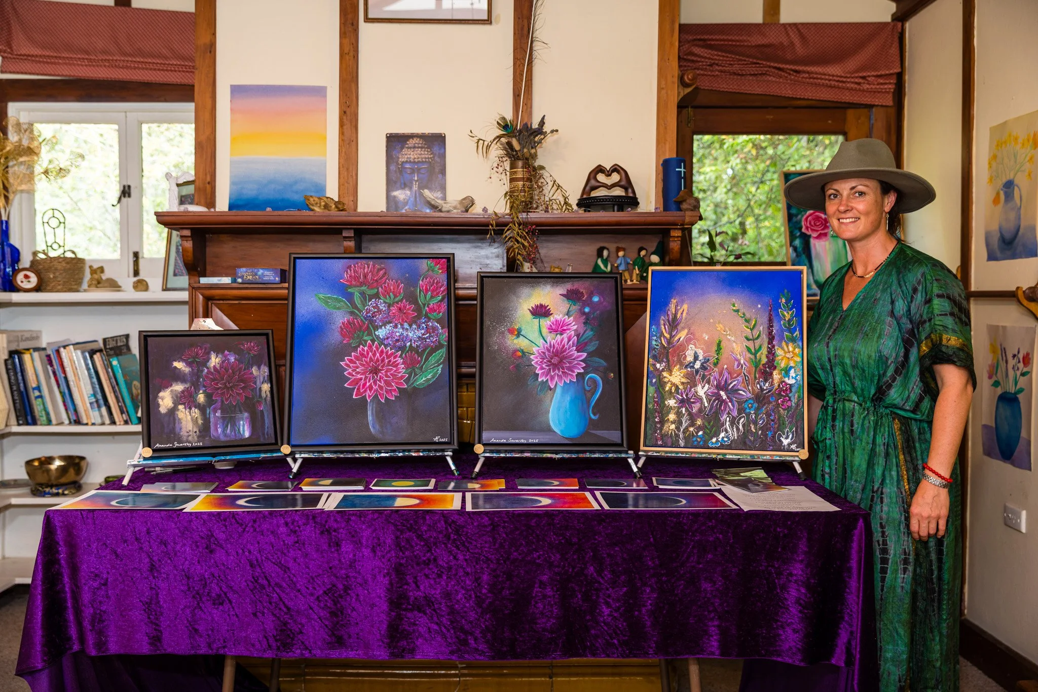Artist woman smiling at her art display with four vibrant floral paintings on easels on a purple table, in a cozy room with books, artwork, and large windows.