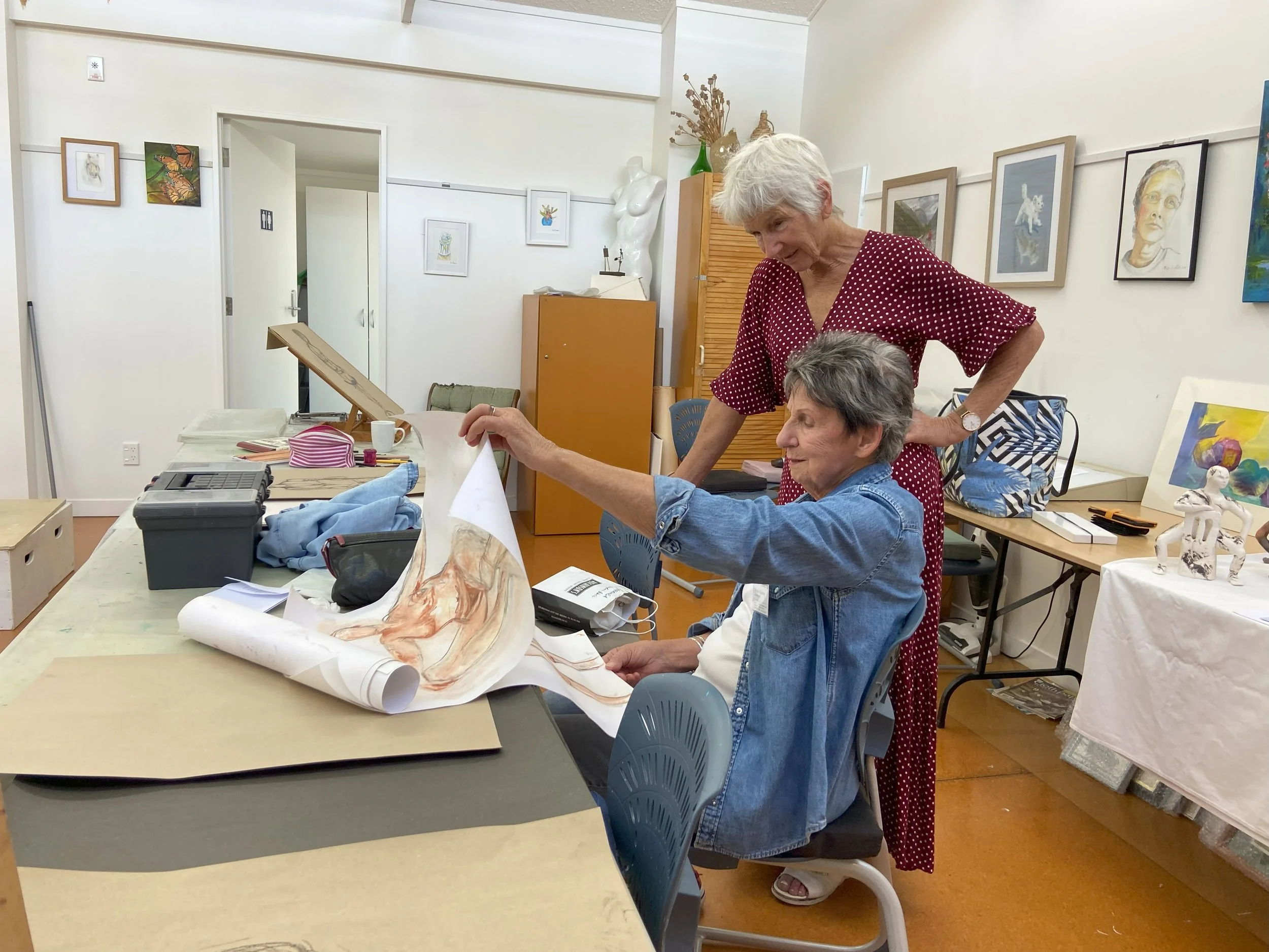 Two women, one older with white hair in a red polka dot dress and the other with short gray hair in a denim jacket, are looking at a large rolled-out artwork on a table in an art studio. The studio has artwork on the walls and various art supplies on the table.