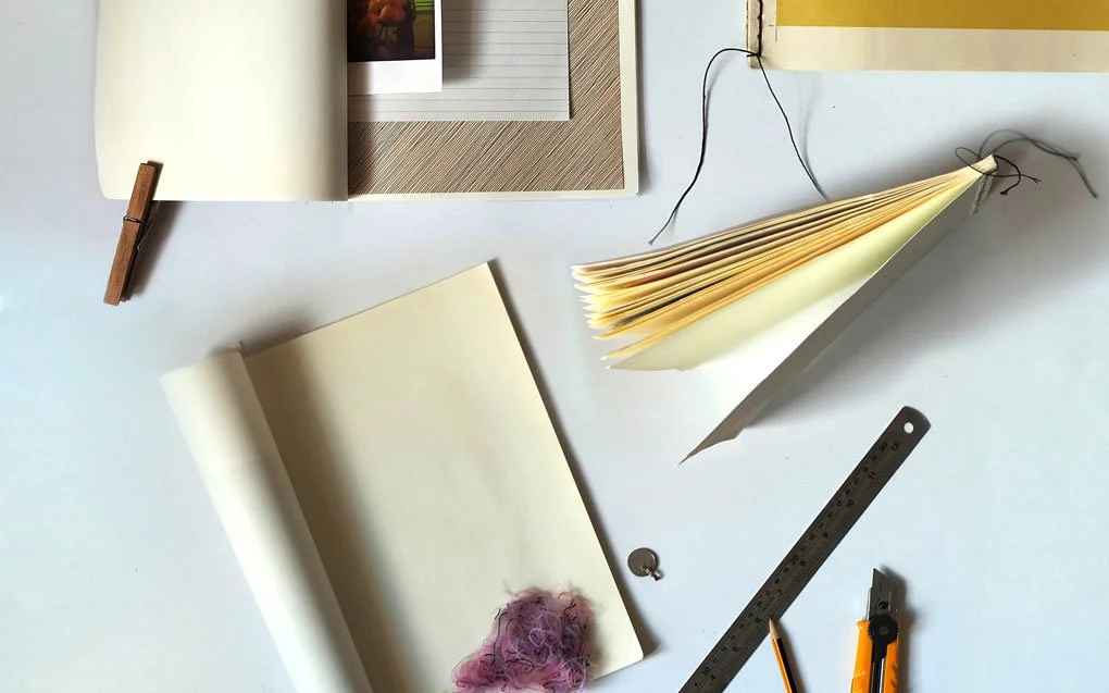 Open sketchbook, closed notebook, pile of yellowed paper, metal ruler, orange utility knife, small silver key, pin, felted pink wool, and clip on a white surface.