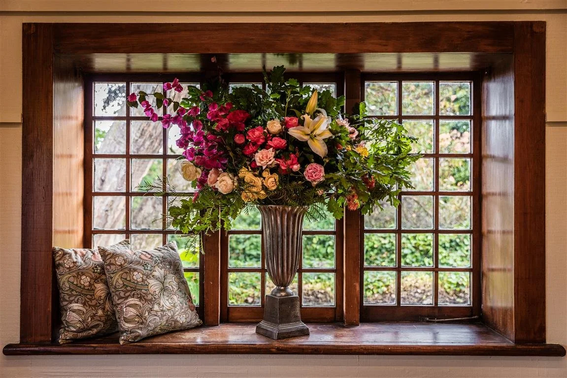 A large flower arrangement in a tall vase on a wooden window sill, with patterned pillows nearby, in front of a multi-pane window showing trees outside.