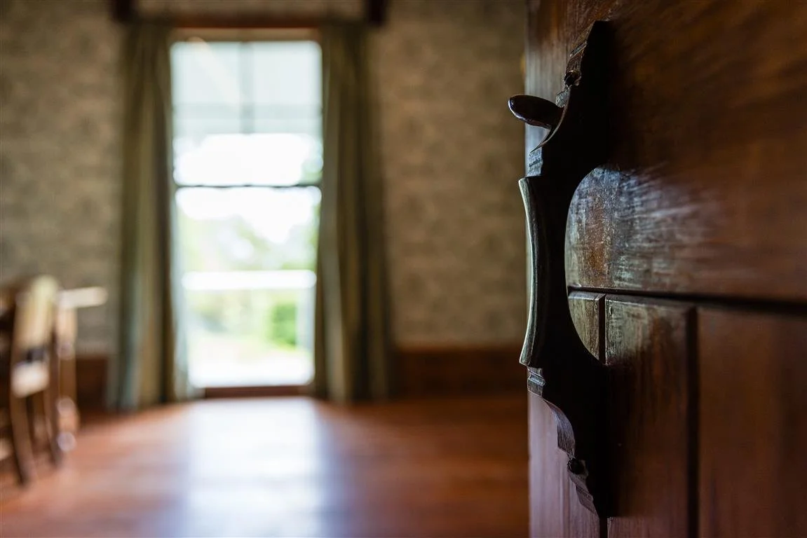 Close-up of a dark wooden door handle on a wooden door, with a blurred background of a room and a window letting in natural light.