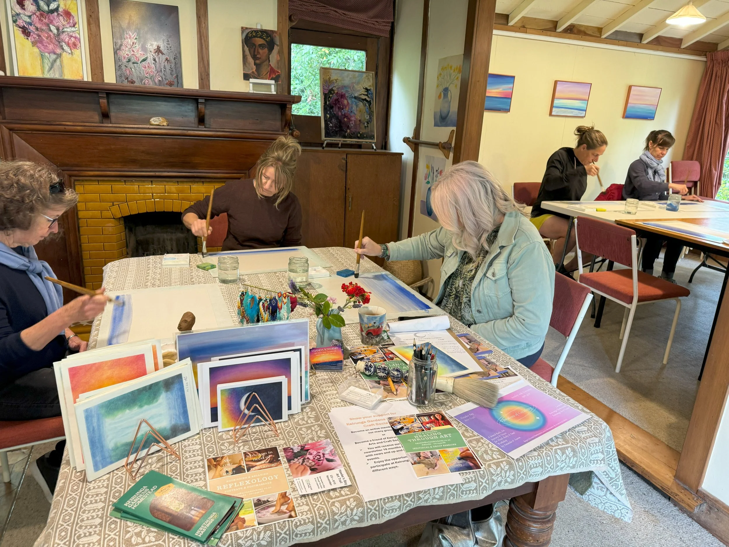 Six women sit at a table, painting and creating art. The table has art supplies, books, and pictures. They are in a cozy room with artworks on the walls and a window showing greenery outside.