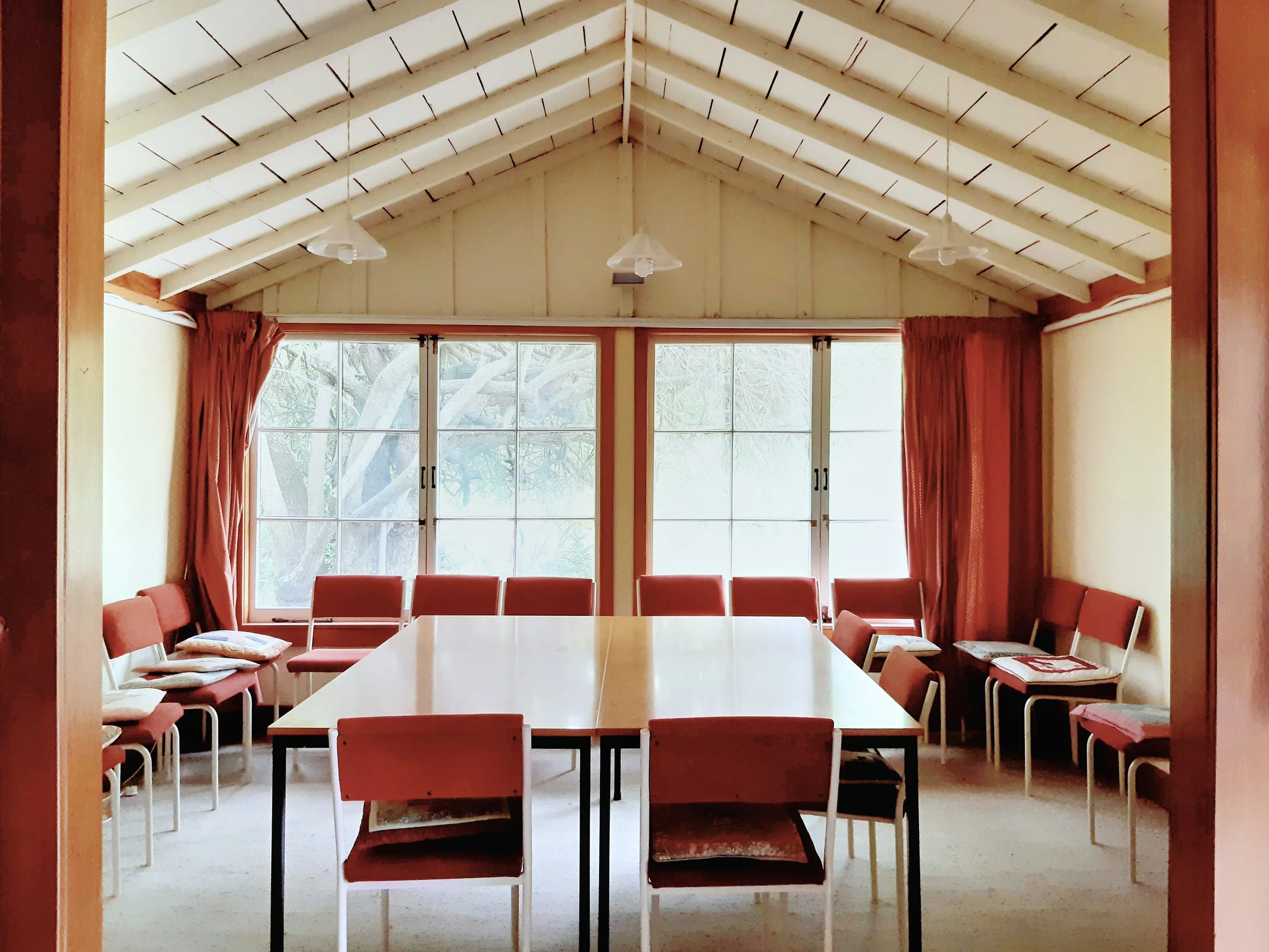 A meeting room with a large rectangular table surrounded by red chairs, a ceiling with exposed beams, large windows with curtains, and trees visible outside.