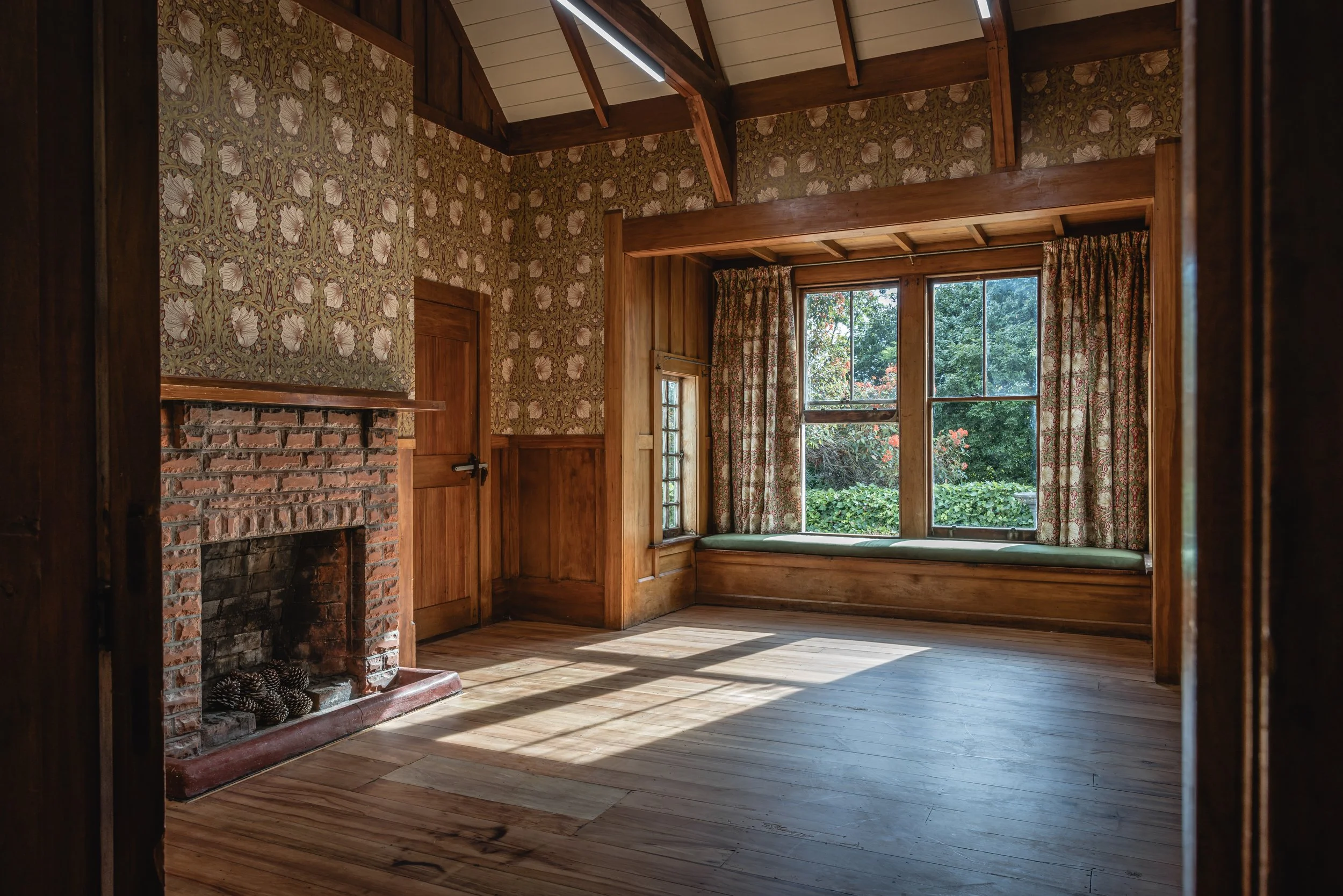 A cozy room with wooden walls, large windows with floral curtains, a brick fireplace, and a window bench with a green cushion, sunlight streaming through the windows.