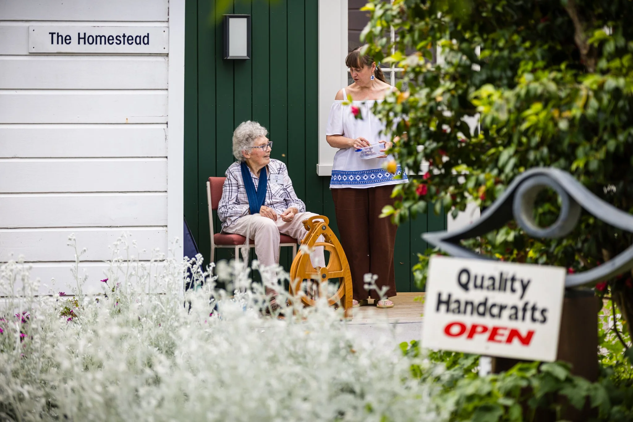 An elderly woman sitting on a porch with a walker nearby, engaged in conversation with a woman standing in front of her holding a brochure, in front of a building labeled 'The Homestead.' There is a sign in the foreground that reads 'Quality Handcrafts OPEN,' and lush greenery and flowers surround the scene.
