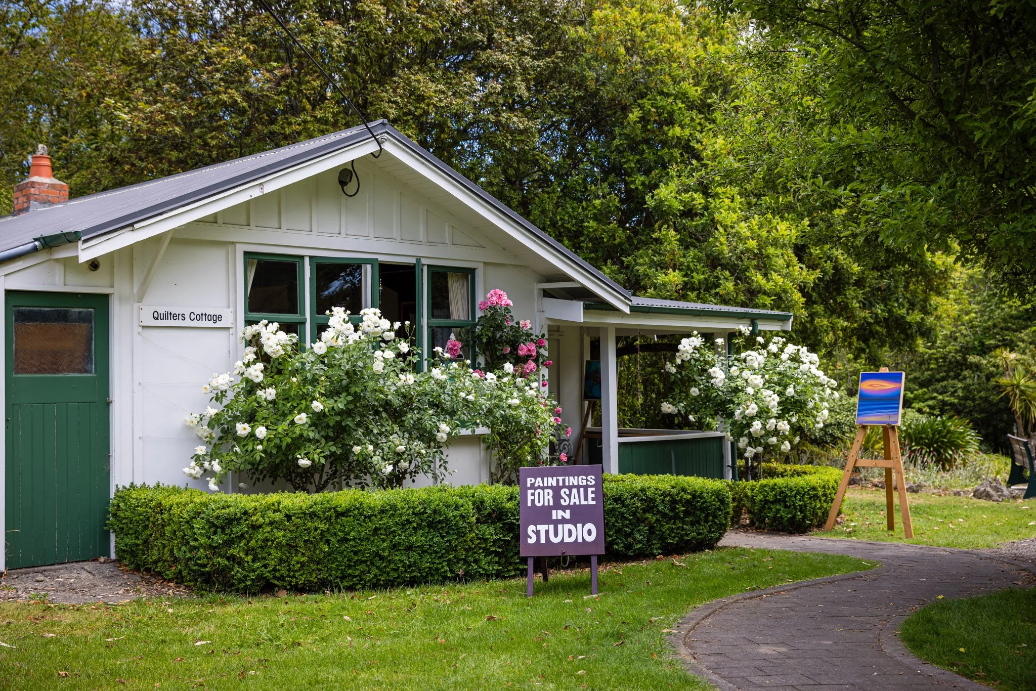 A white cottage with green trim named Quilters Cottage, surrounded by blooming white and pink roses and trimmed bushes. A sign in front advertises paintings for sale in the studio, and an easel with an abstract painting is visible to the right of the house. A curved pathway leads to the house among greenery and trees.