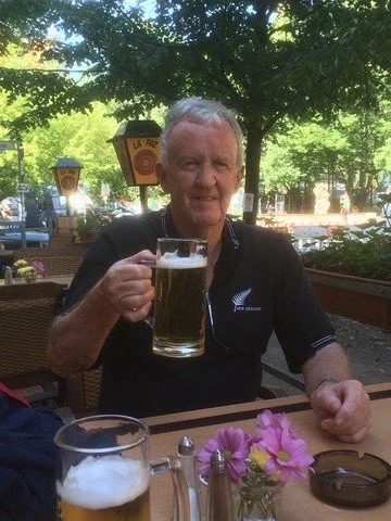 An elderly man sitting outdoors at a table, holding a mug of beer, with pink flowers and a glass of beer on the table in front of him, under a tree with green leaves.
