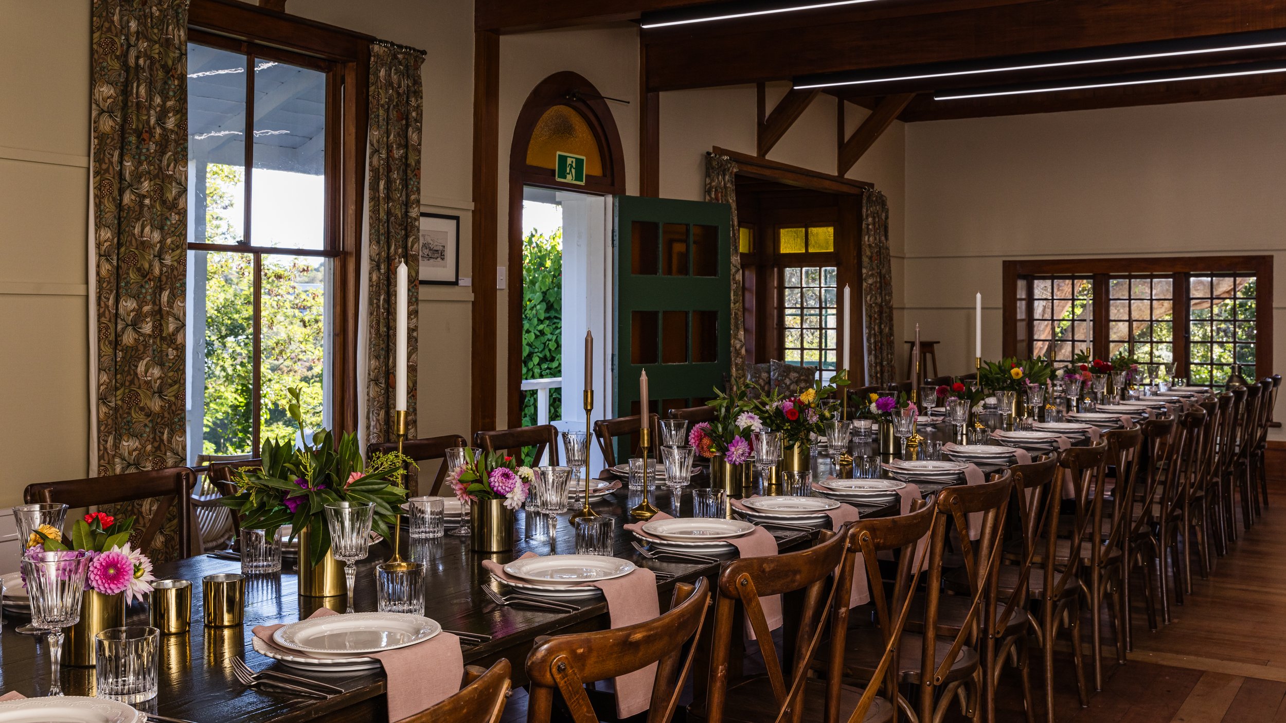 Long wooden dining table set for a formal event with plates, glasses, pink napkins, and floral centerpieces in gold vases, in a rustic room with large windows and wooden beams.
