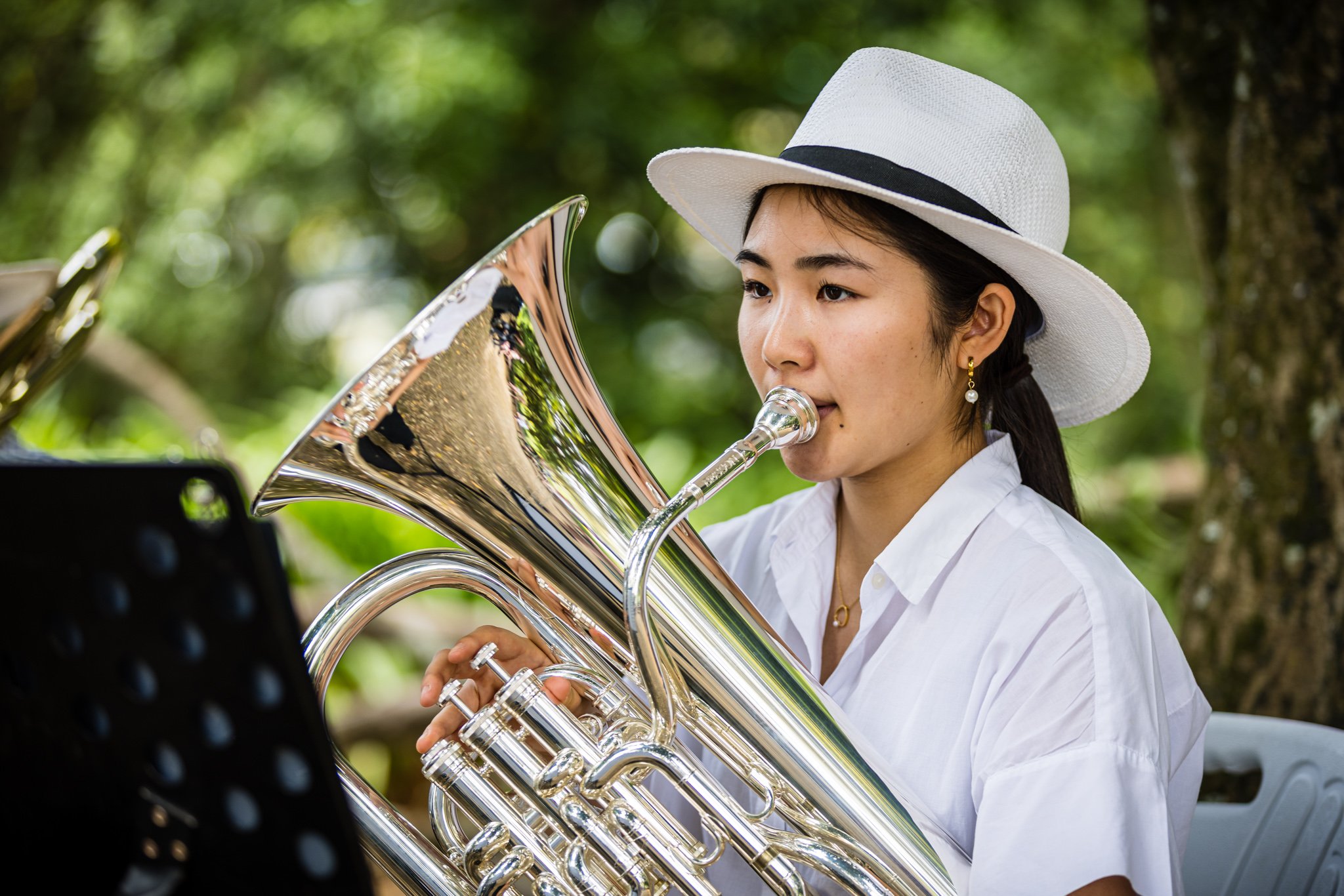 Young woman playing a silver euphonium outdoors, wearing a white hat and white shirt with a black band on her hat, in a lush green setting.