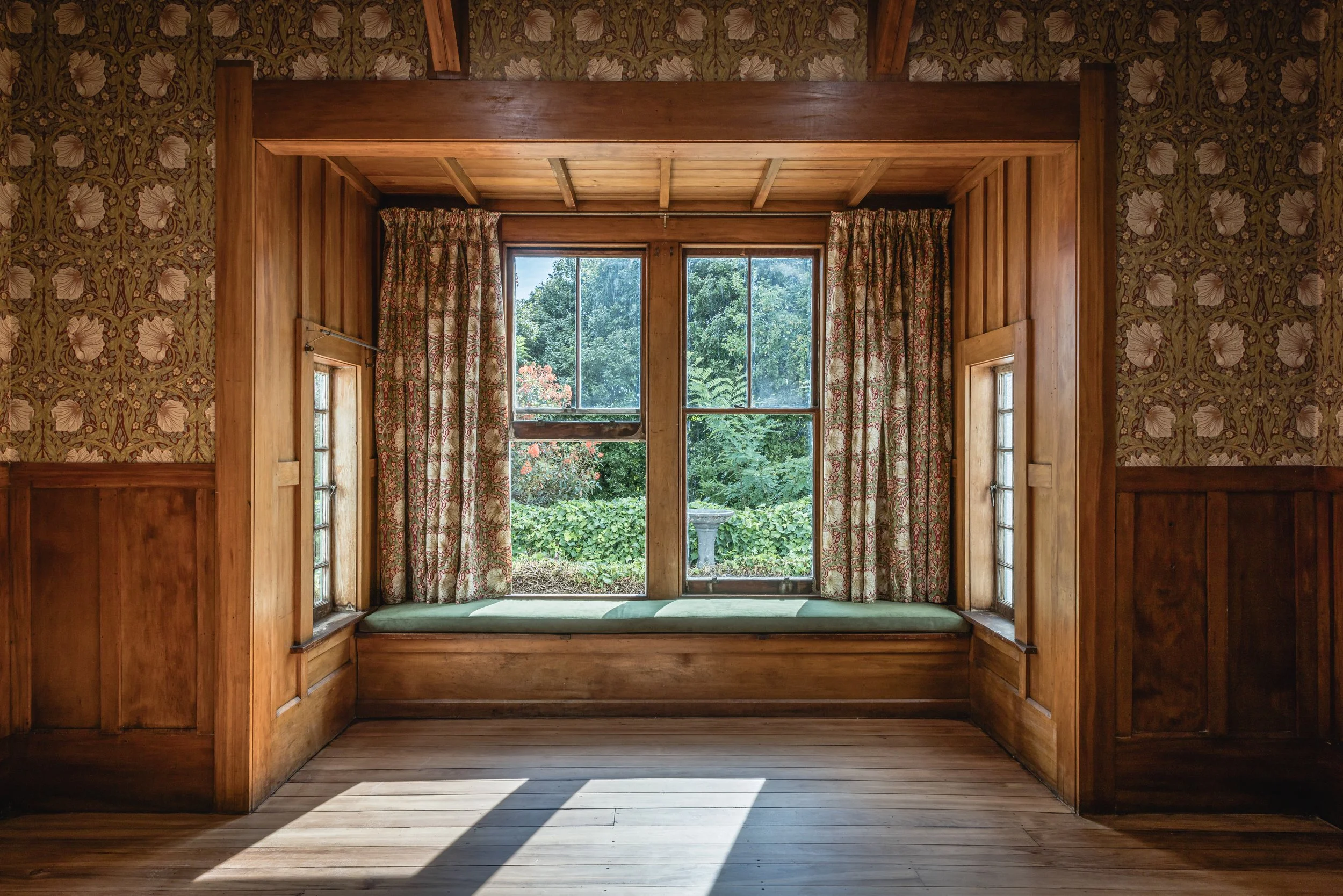 A cozy wooden window nook with a green cushioned seat, framed by floral curtains and surrounded by wooden paneling, overlooking a lush garden with trees and a birdbath.
