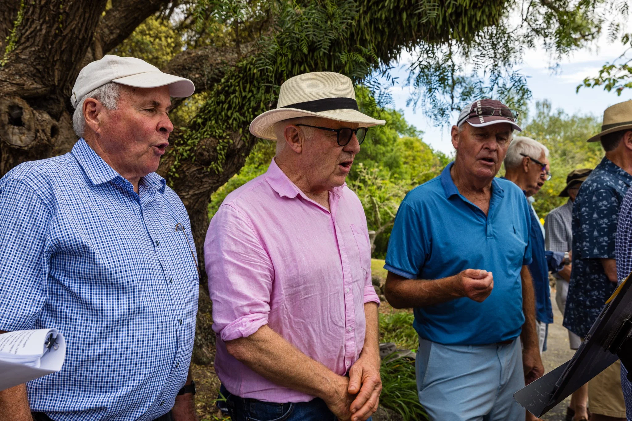 A group of elderly men singing outdoors, dressed in casual summer clothes with hats, with trees and greenery behind them.