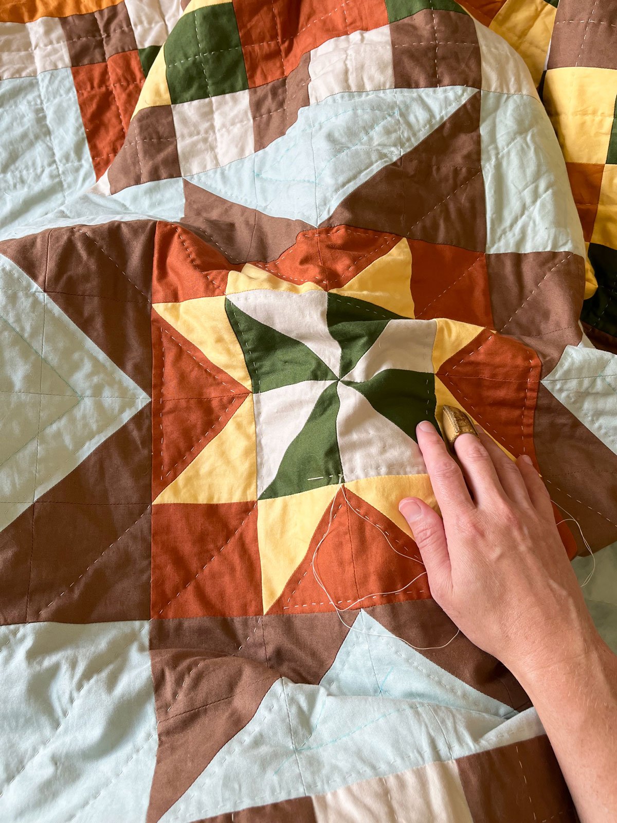 Close-up of a colorful quilt with a patchwork design of squares and triangles in earth tones, with a person's hand holding the fabric.