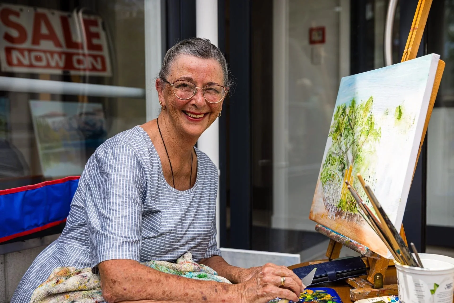 An elderly woman with glasses smiling while painting outdoors, sitting at an easel with a colorful landscape painting of trees and sky, surrounded by art supplies.