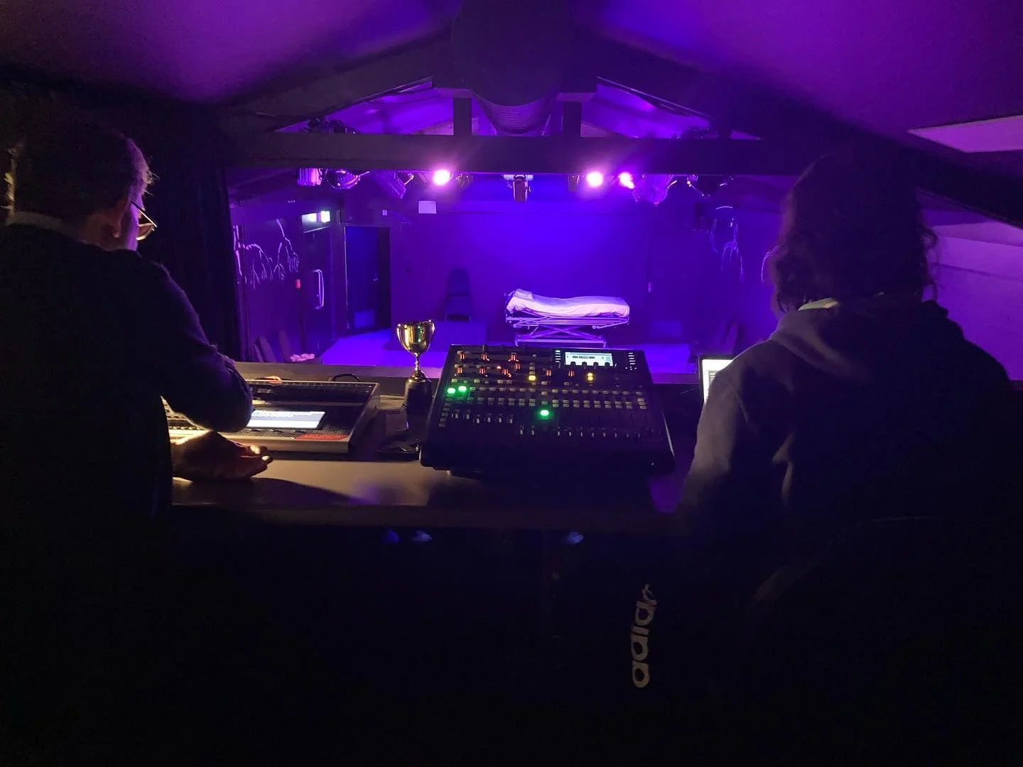 View of a dimly lit theater stage with purple lighting, seen from behind a control booth with two audio engineers working at mixing consoles.