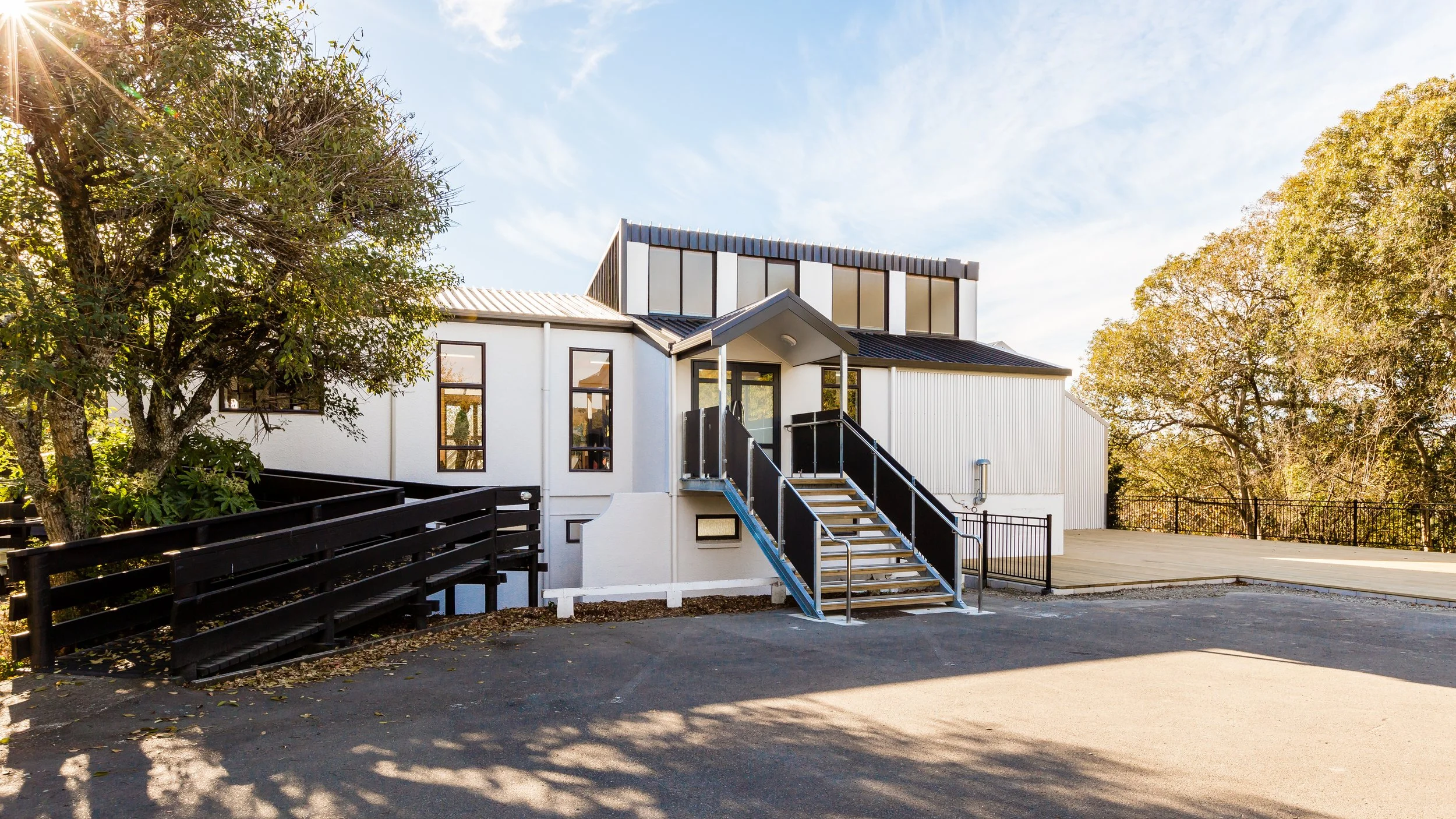 Modern white building with black trim and stairs leading up to the entrance, surrounded by trees and a spacious parking lot.