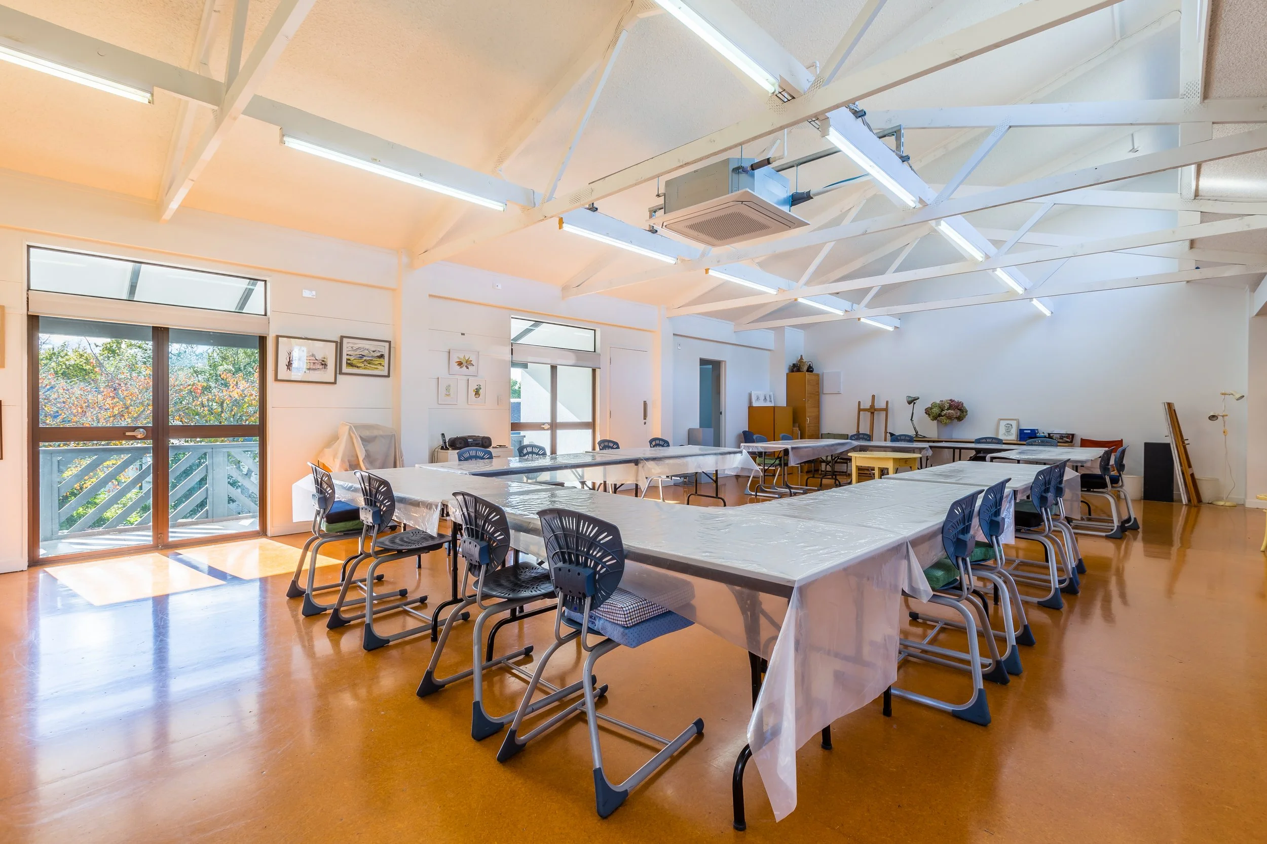 A spacious room set up for a group activity with 'U'-shaped long tables covered in disposable tablecloths, surrounded by black plastic chairs, with artwork on the walls and large windows letting in natural light.