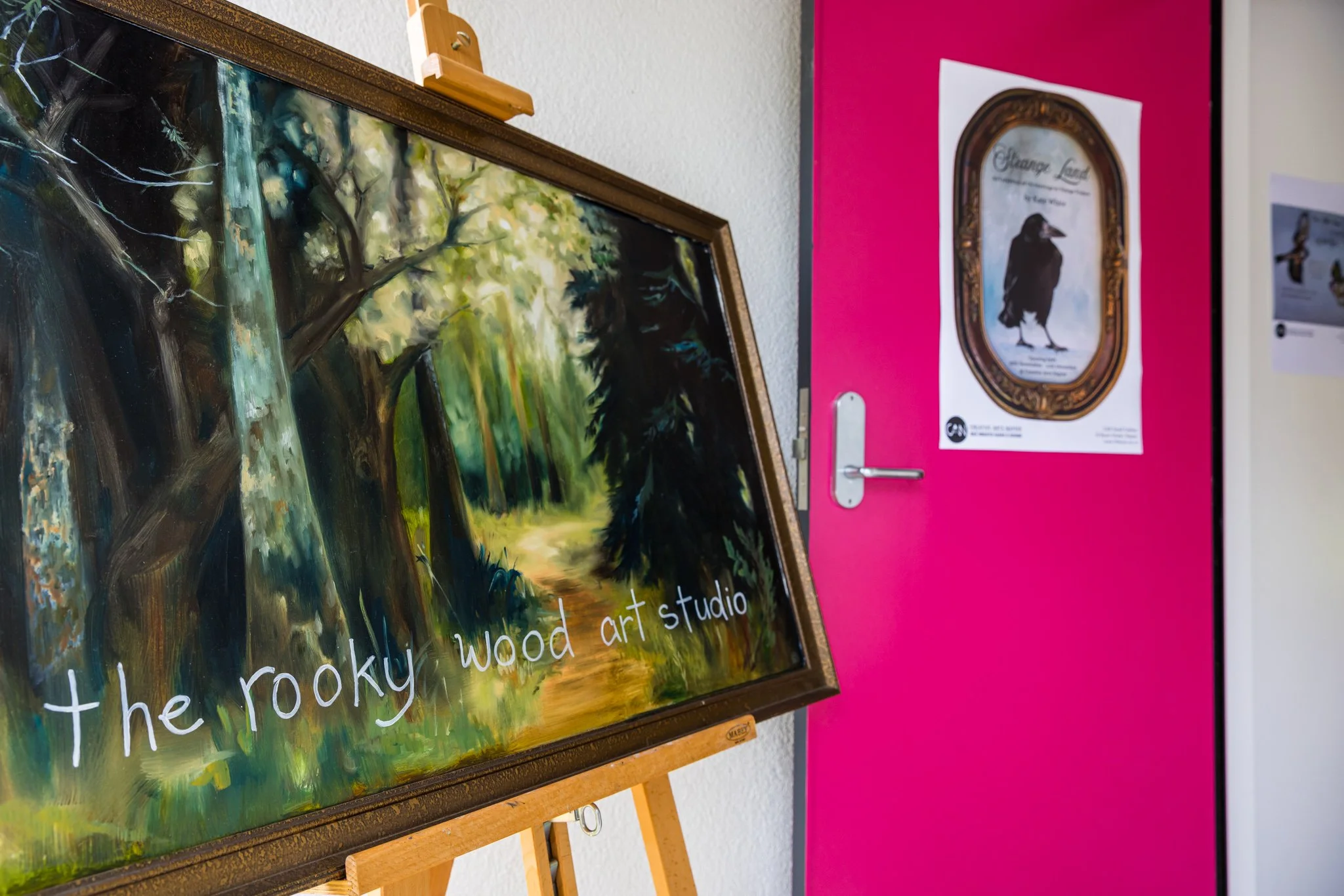 A framed painting of a forest scene with green trees and a bright path, leaning against an easel on a white wall. Part of a pink door with a poster of a bird inside a decorative frame is visible in the background.