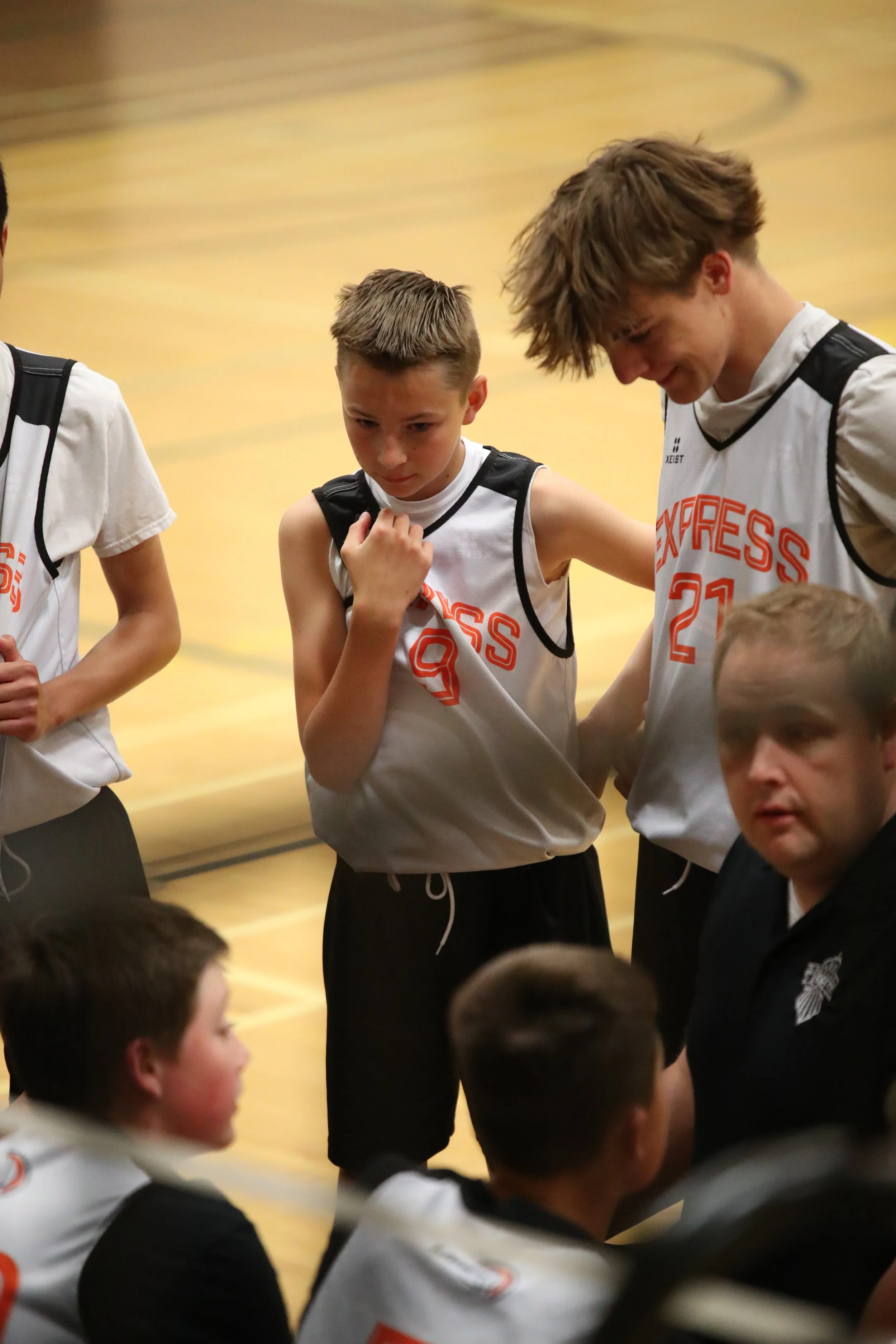 A group of young basketball players in white jerseys with orange lettering, sitting in a gymnasium during a timeout.