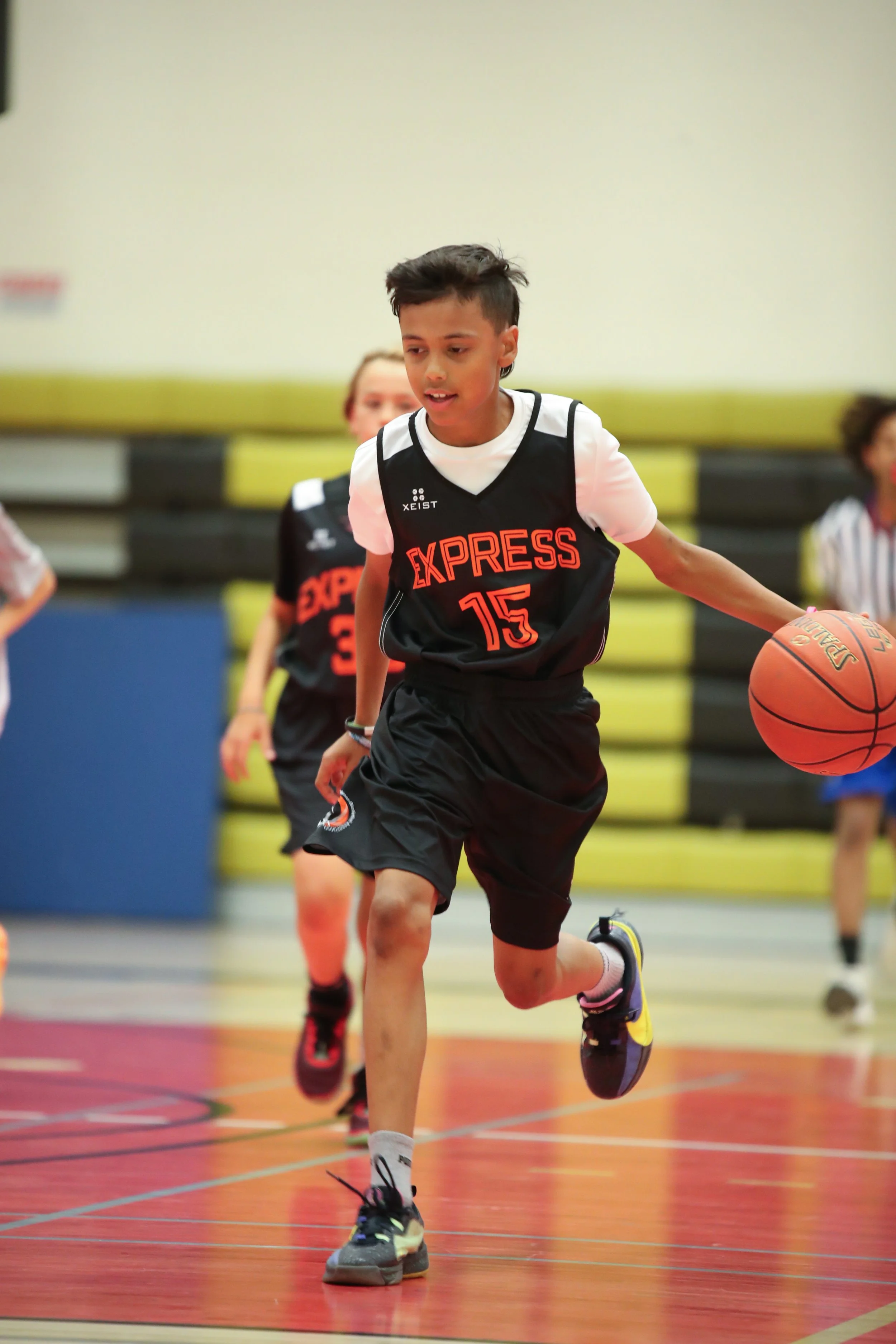 Young boy playing basketball in a gymnasium, dribbling the ball, wearing a black jersey with 'EXPRESS' and number 15.