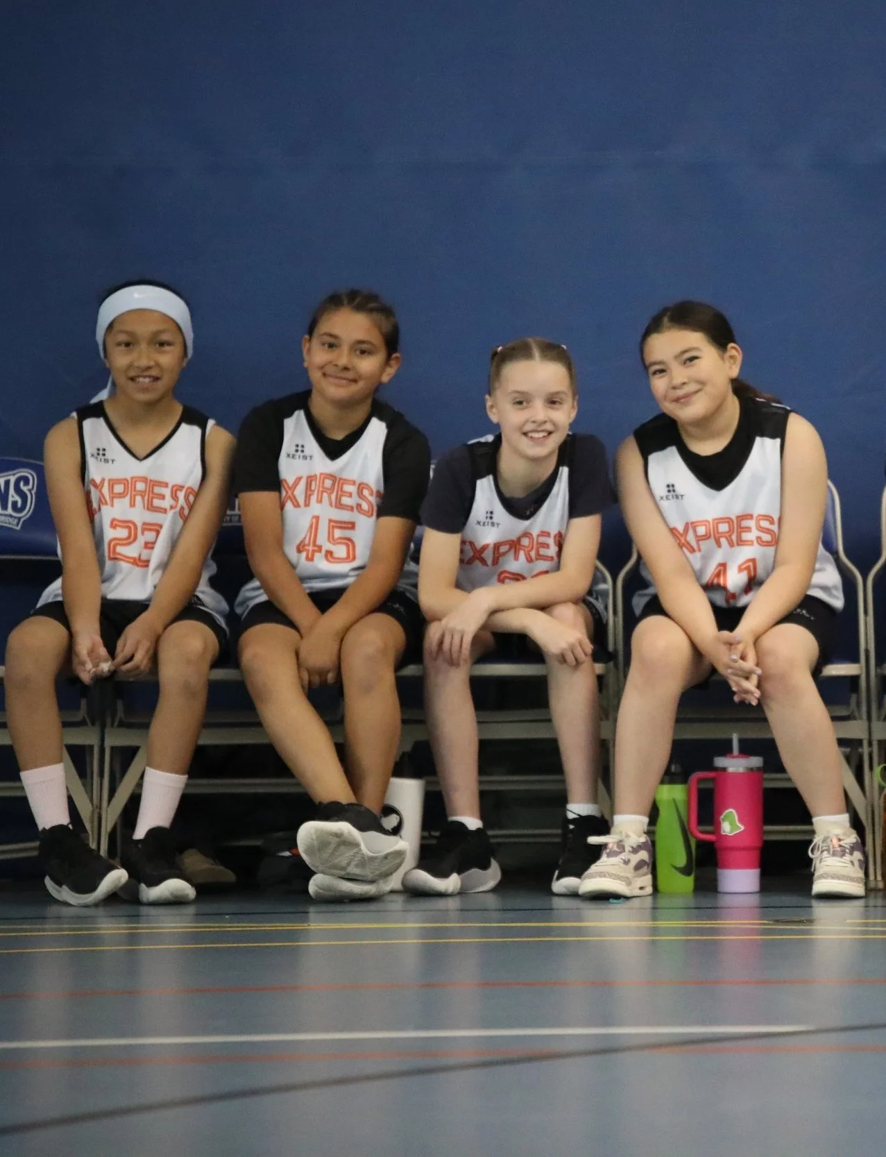 Four young girls sitting on a bench in a gymnasium, wearing basketball uniforms with 'EXPRESS' written on them, smiling at the camera.