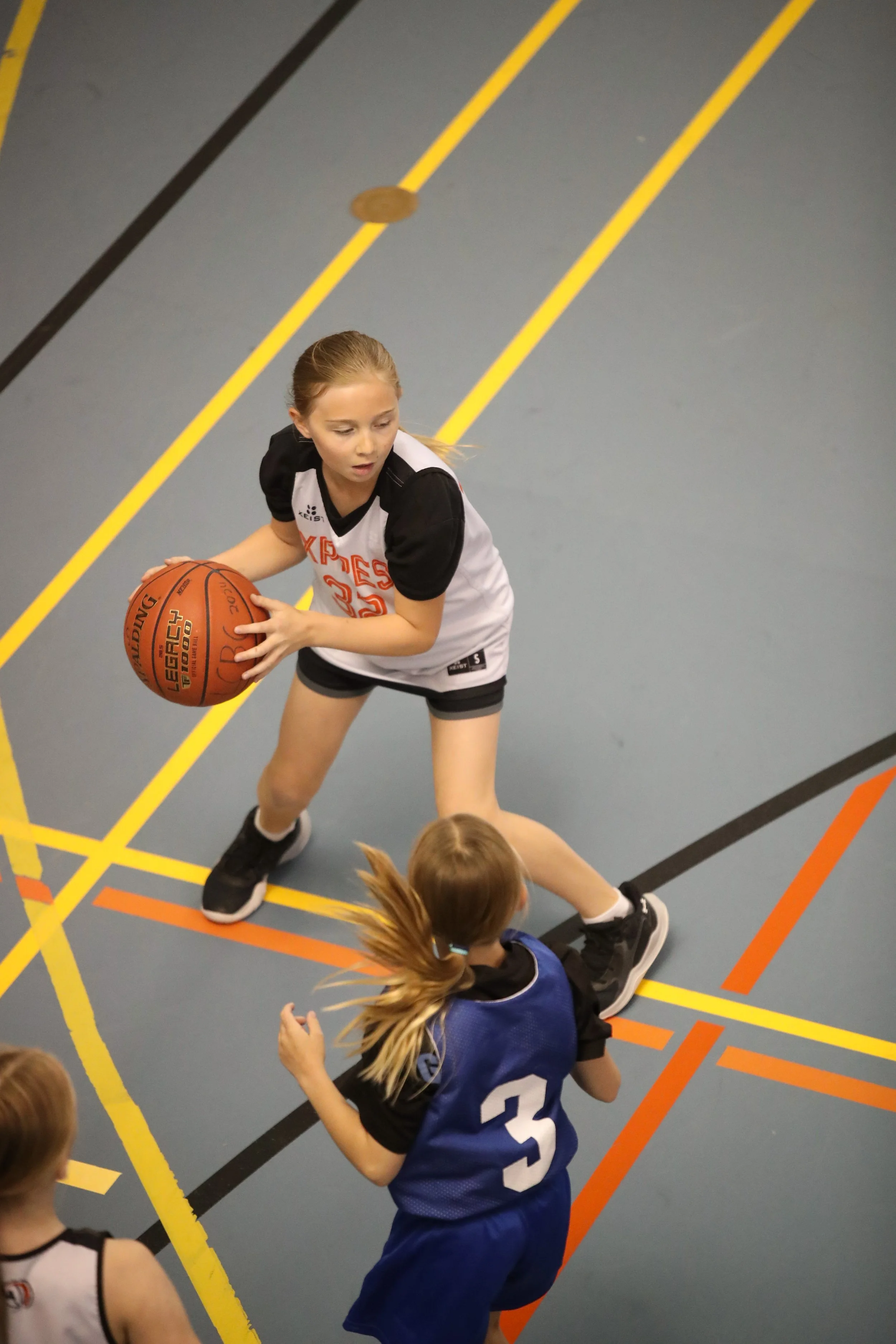 A young girl with a basketball in her hands, wearing a black and white sports uniform, is playing basketball in an indoor gym.