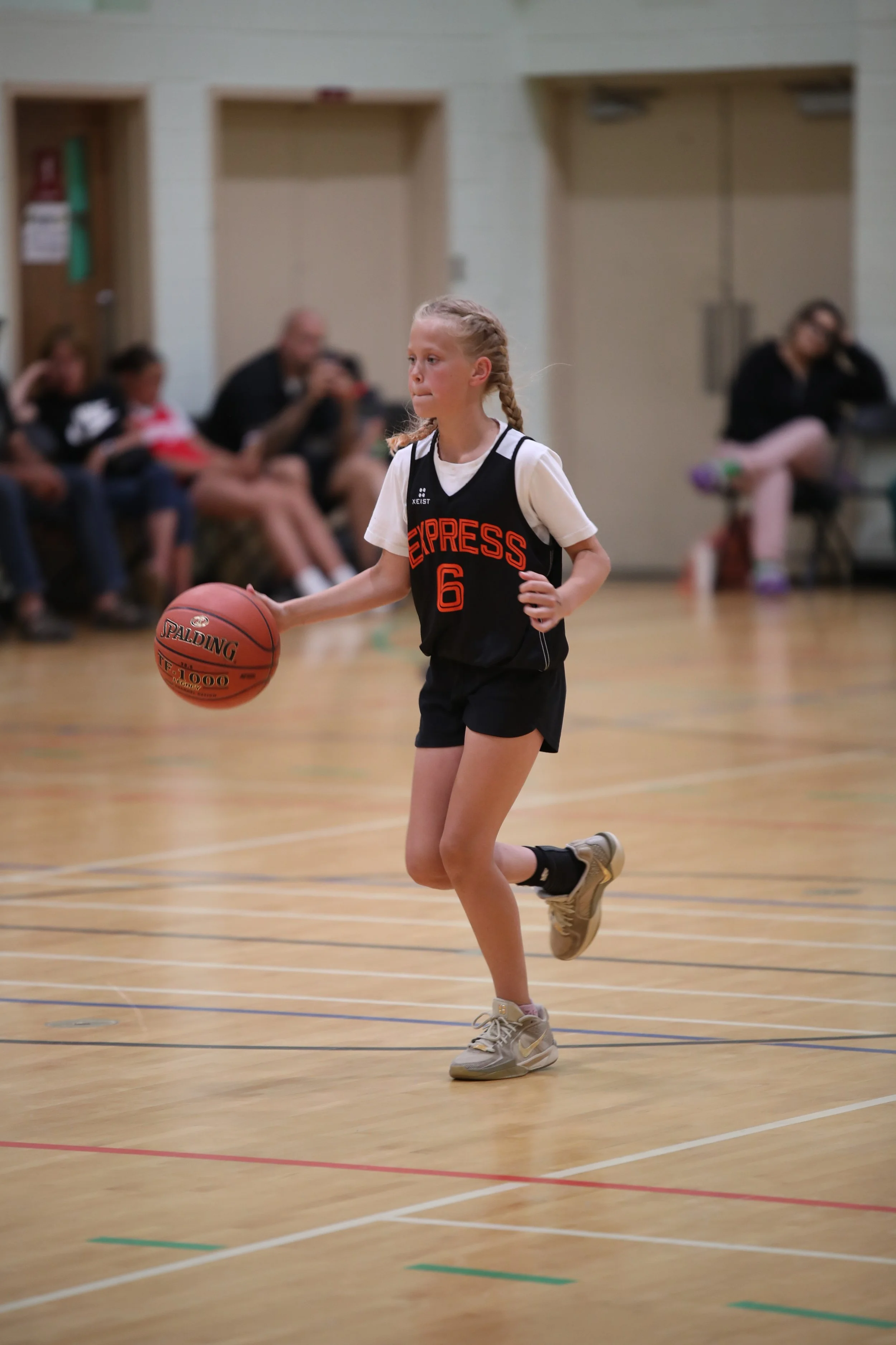 A young girl playing basketball on an indoor court, dribbling a Spalding basketball, wearing a black and white sports uniform with the number 6 and the word 'EXPRESS' on it.