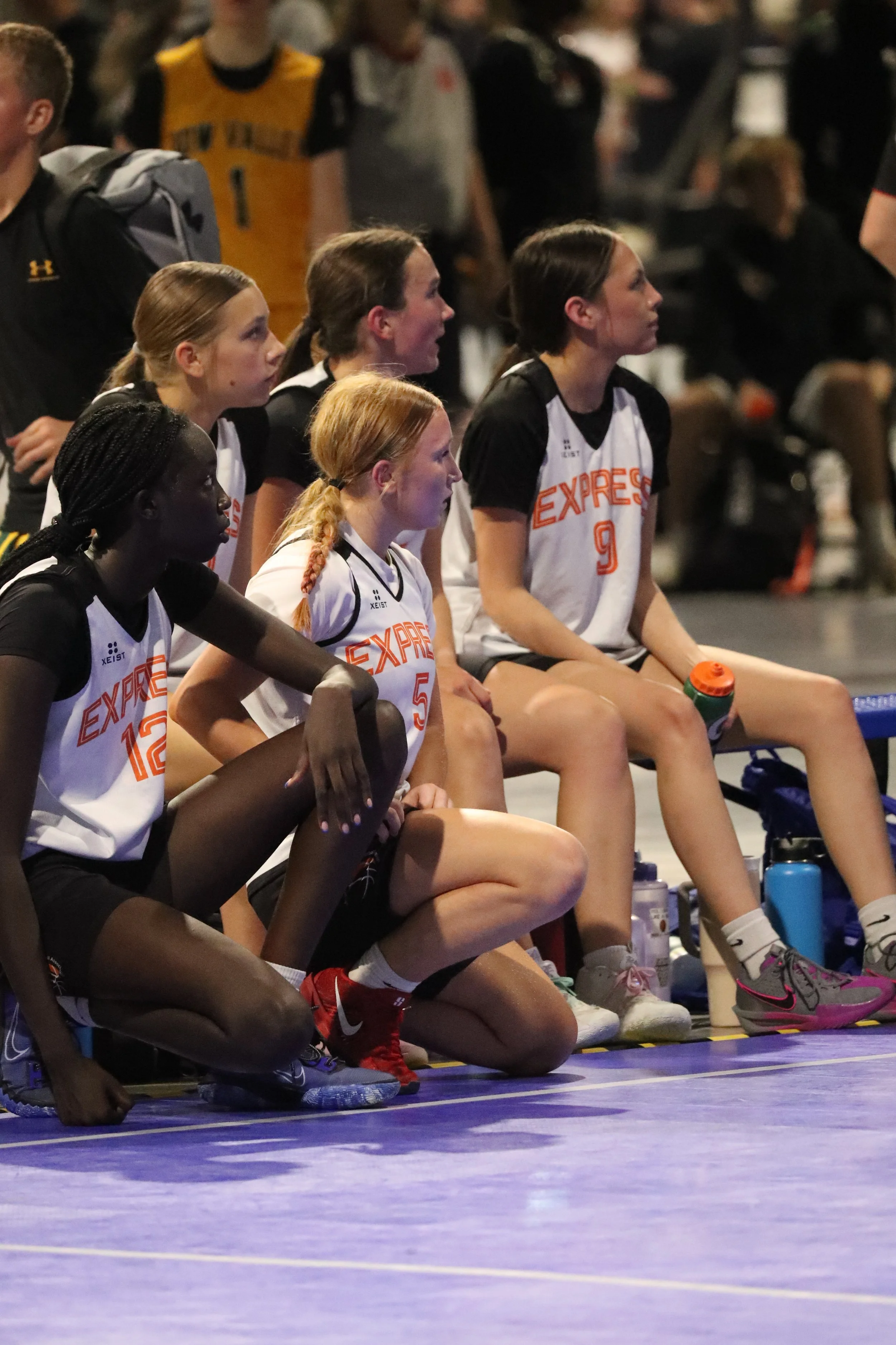 A women's basketball team sitting on the bench during a game, wearing white jerseys with 'EXPRESS' and numbers, on an indoor court.