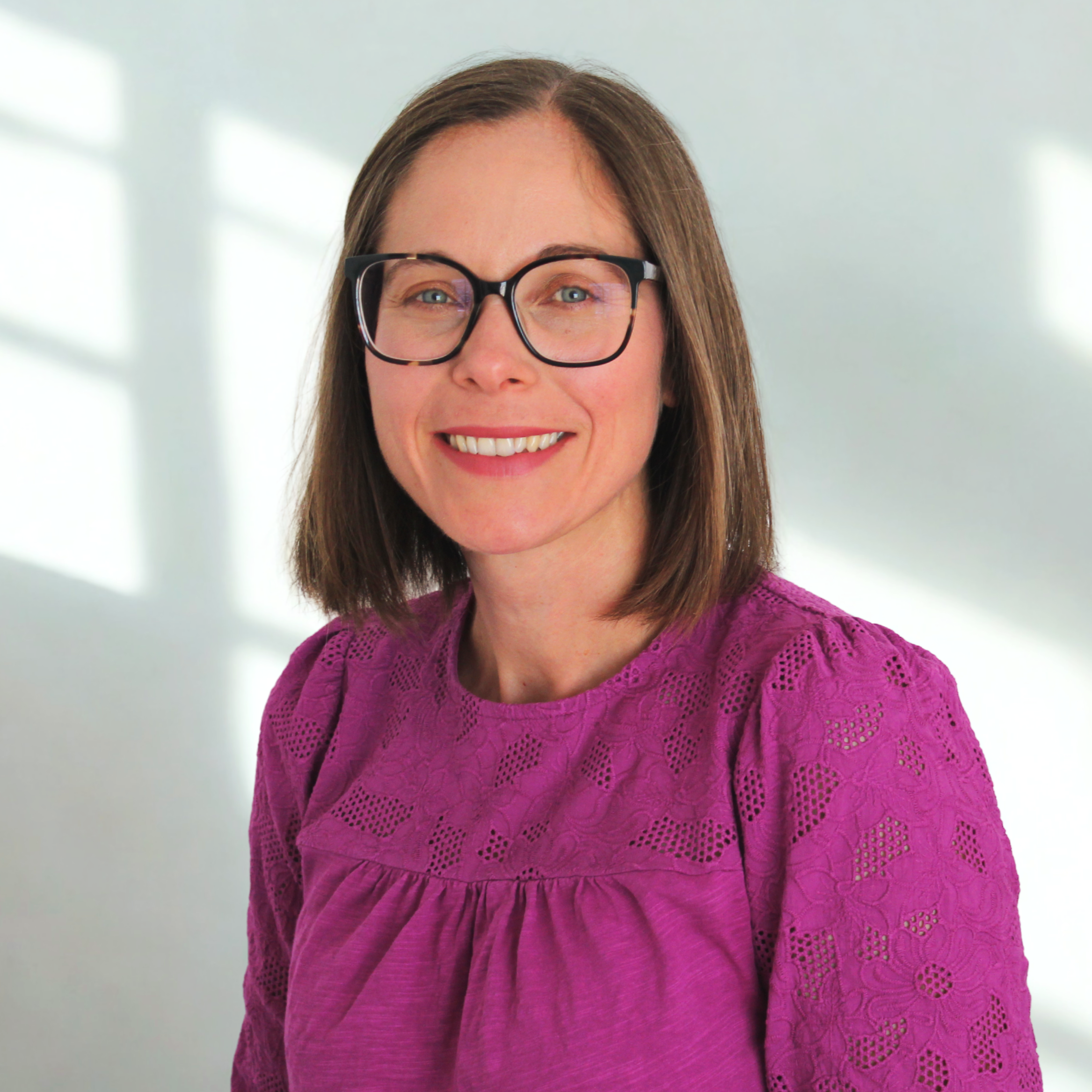 A woman with shoulder-length brown hair and black-rimmed glasses smiling at the camera, wearing a purple lace top, standing in a bright room with blurred window light in the background.