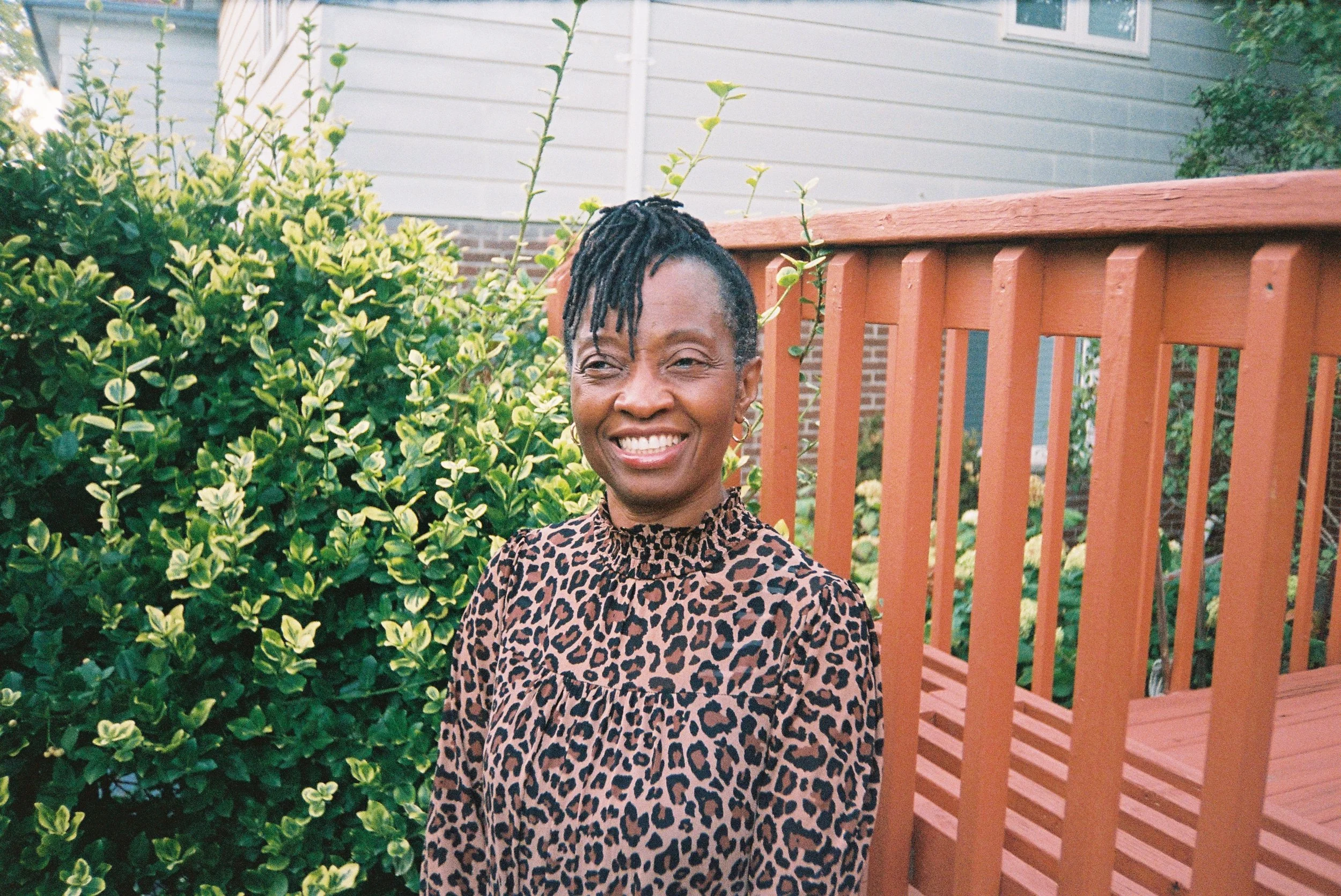 A woman with short, curly hair styled in dreadlocks tied into a top bun, smiling while standing outdoors near lush green bushes and a wooden fence, with a house in the background.