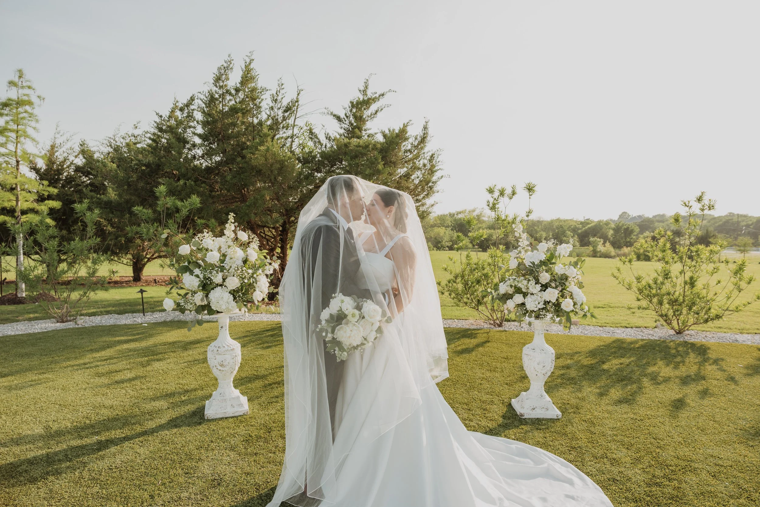 Bride and groom kissing under a veil at an outdoor lawn wedding ceremony framed by white floral arrangements and greenery coordinated by Ignited Events in Dallas Fort Worth