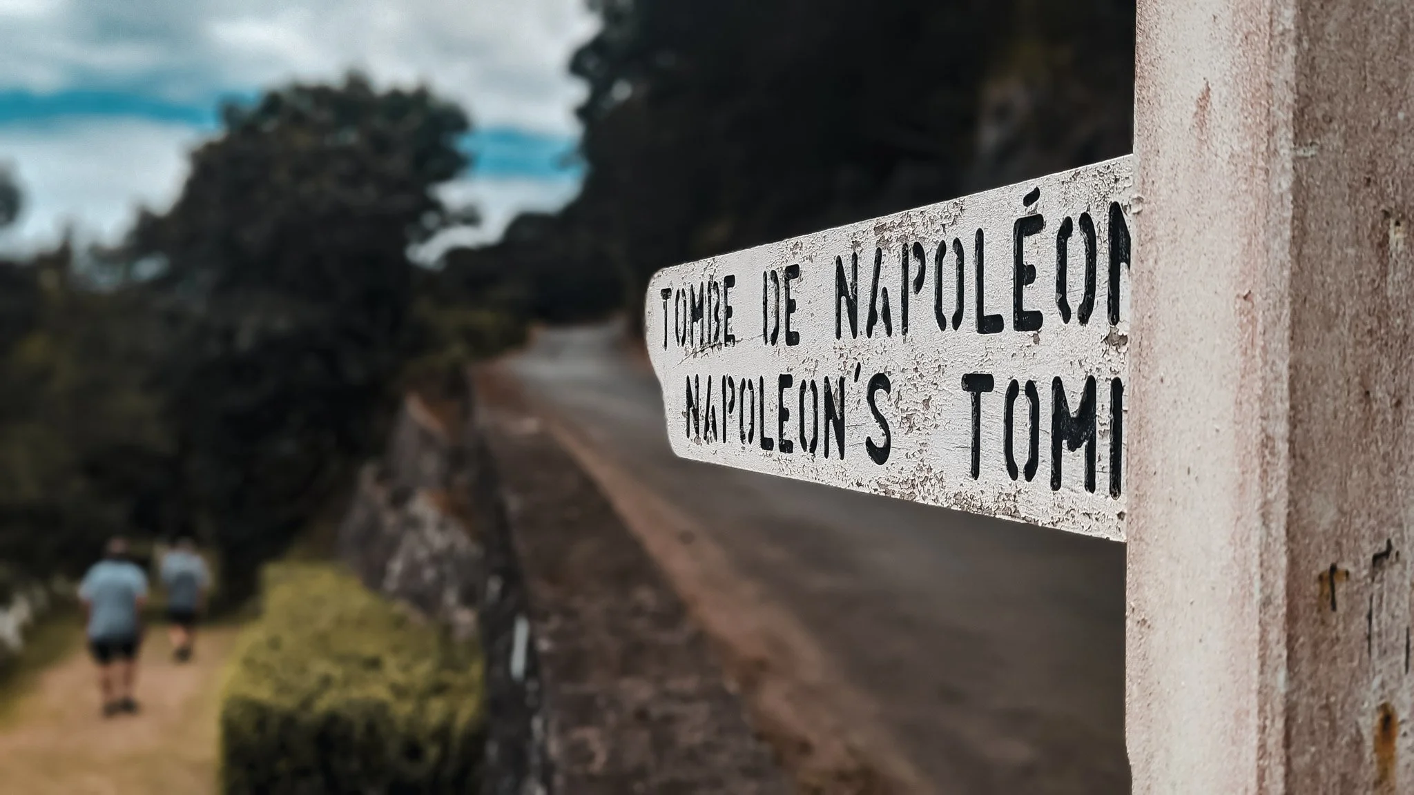 Close-up of a sign reading, "TOMBE DE NAPOLÉON NAPOLEON'S TOM", attached to a concrete wall, with two hikers walking along a dirt path in a scenic outdoor setting in the background.